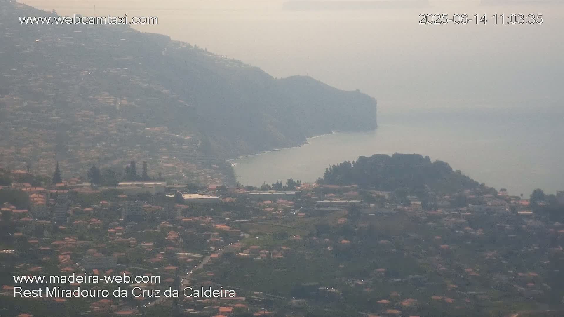 A hazy coastal town nestled against a steep hillside overlooking a calm ocean.