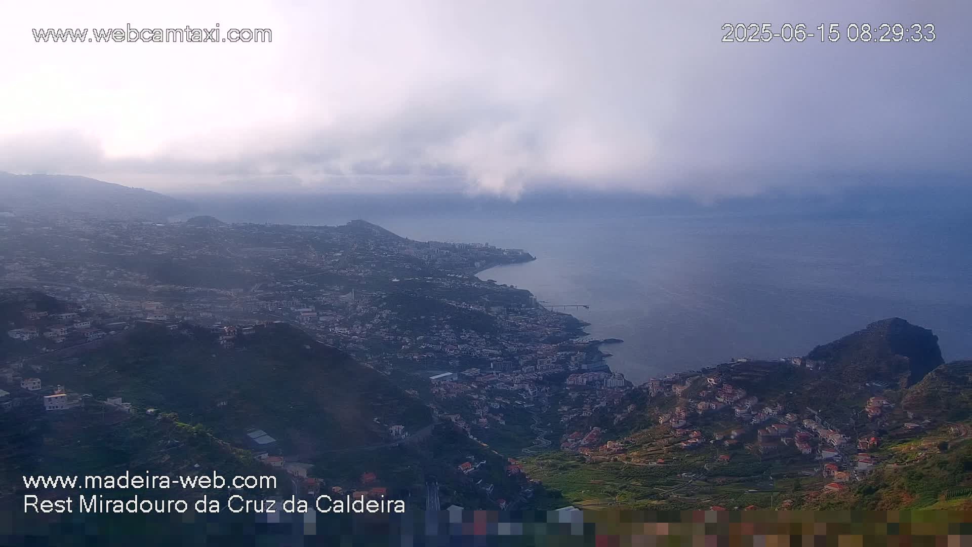 A hazy coastal city sprawls along a hillside overlooking a calm ocean under a cloudy sky.