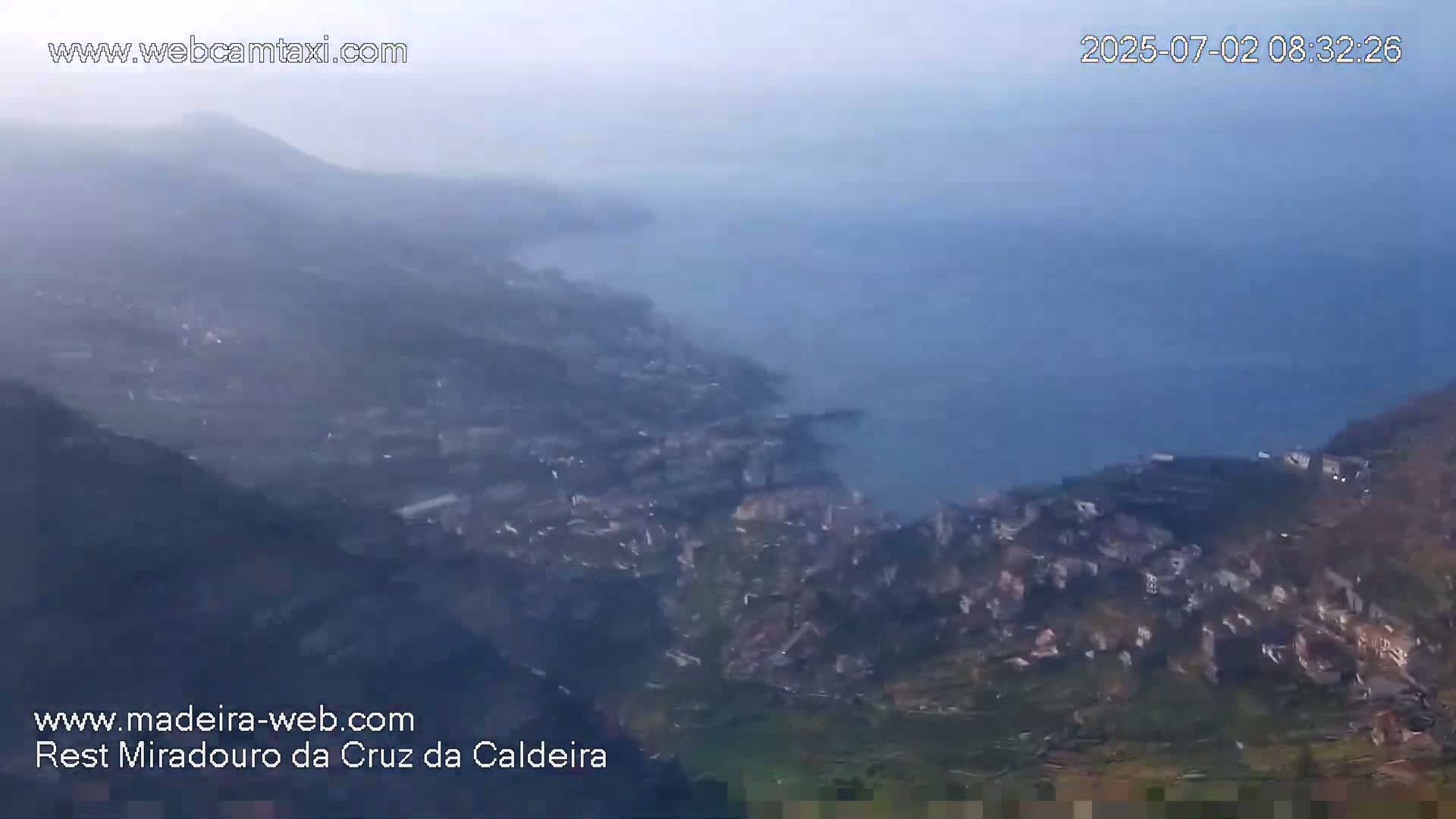 A hazy, high-angle view shows a coastal town nestled into a hillside, with a body of water and mountains visible in the distance.