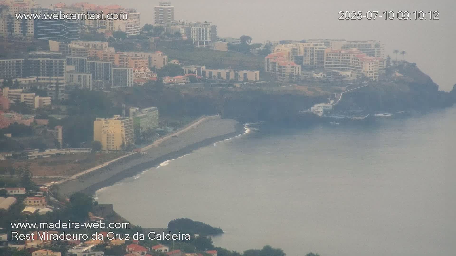A hazy coastal view shows a dark-sand beach curving along a rocky shoreline, bordered by numerous buildings and high-rises.