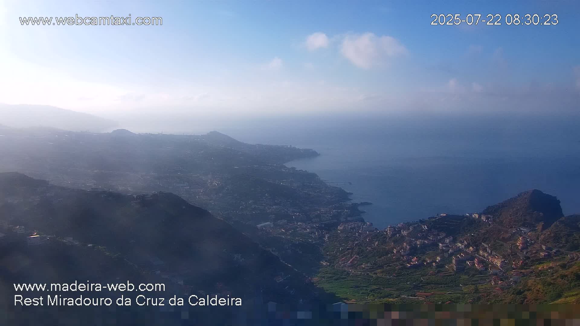 A hazy, high-angle view shows a coastal town nestled in a mountainous region overlooking a calm ocean under a partly cloudy sky.