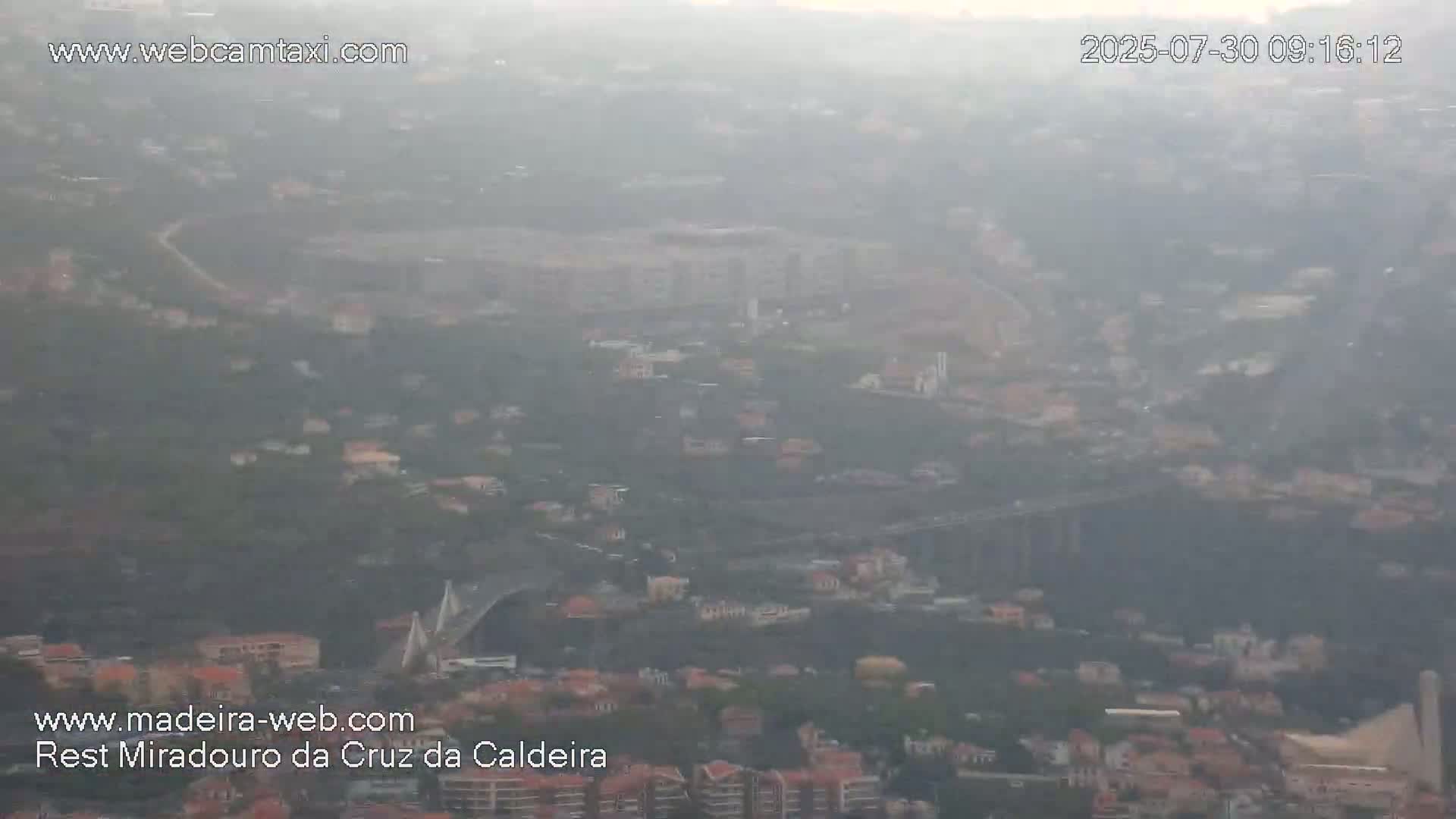 A hazy, long-distance view shows a bridge and a sprawling town nestled among hills.