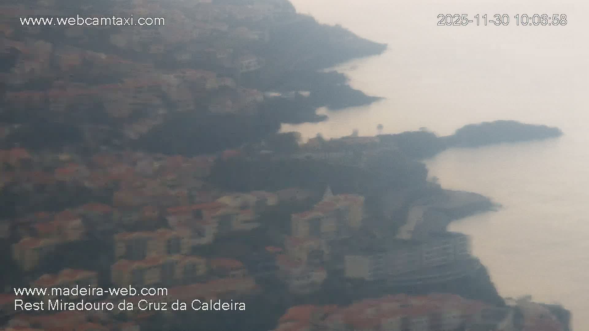 A densely built coastal area featuring numerous buildings on sloping hillsides overlooks a calm sea under heavy haze or fog.