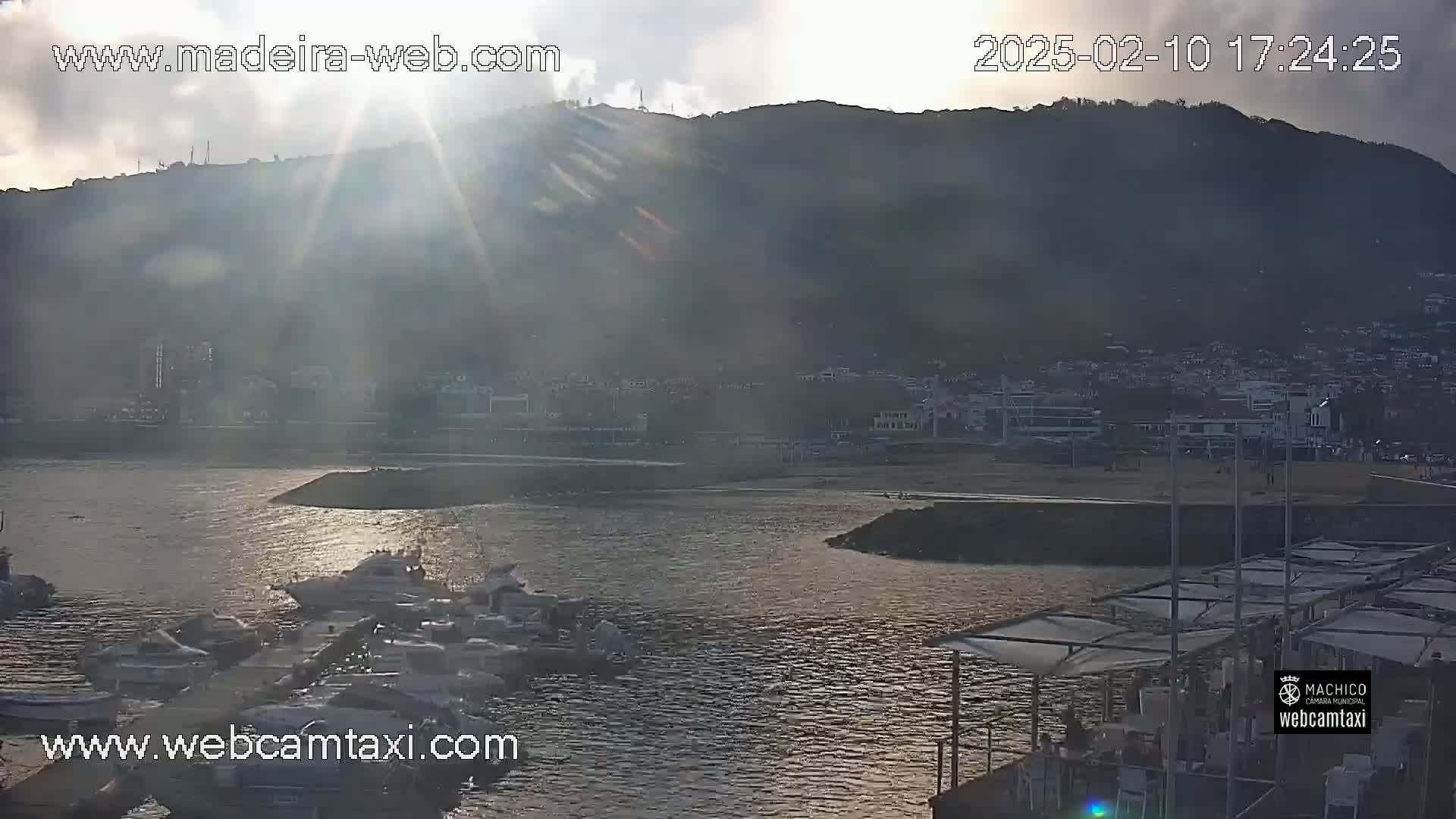 A hazy sunset illuminates a harbor filled with boats, nestled below a hill dotted with buildings.