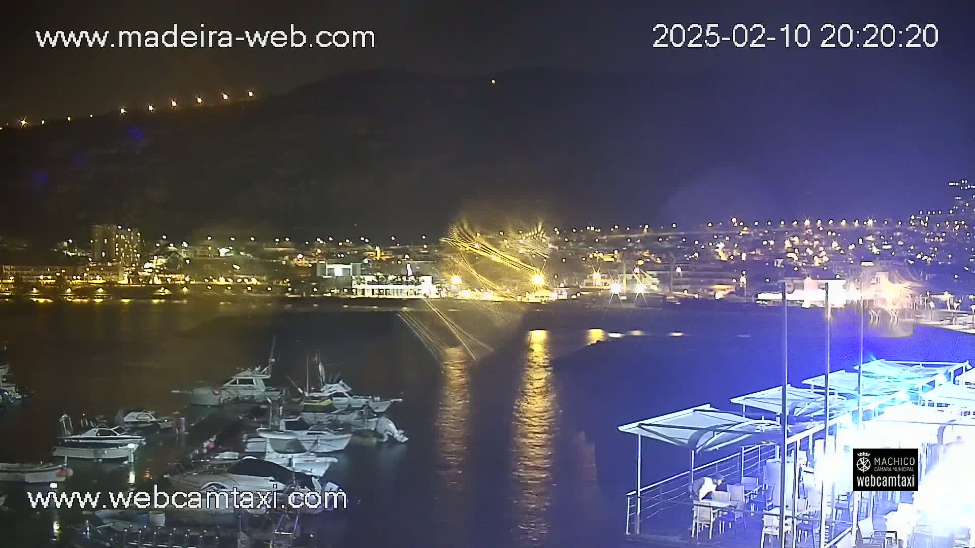 A nighttime view of a harbor filled with boats, with a town illuminated on a hill in the background under a clear, dark sky.