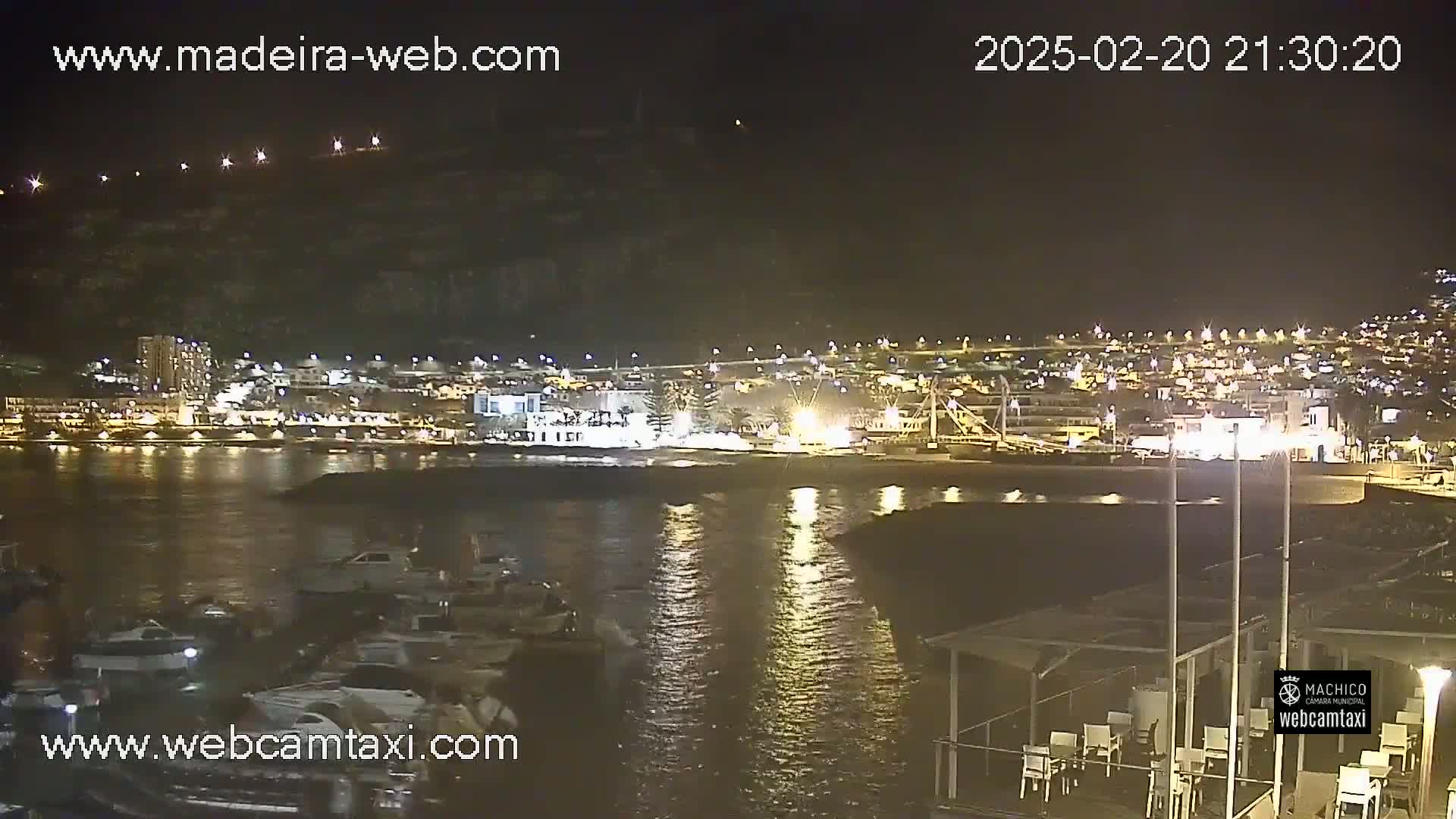A nighttime view of a harbor with boats, a town lit up on the shore, and calm water reflecting the lights.