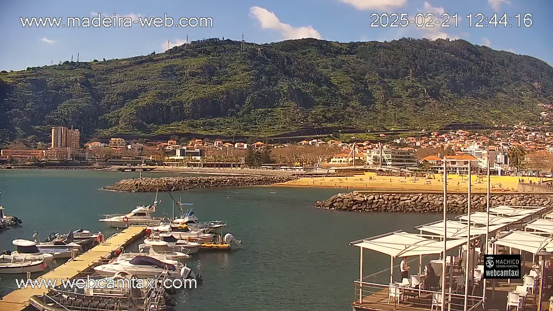 A marina filled with boats sits at the base of a green, mountainous coastline on a sunny day, with a sandy beach visible in the background.