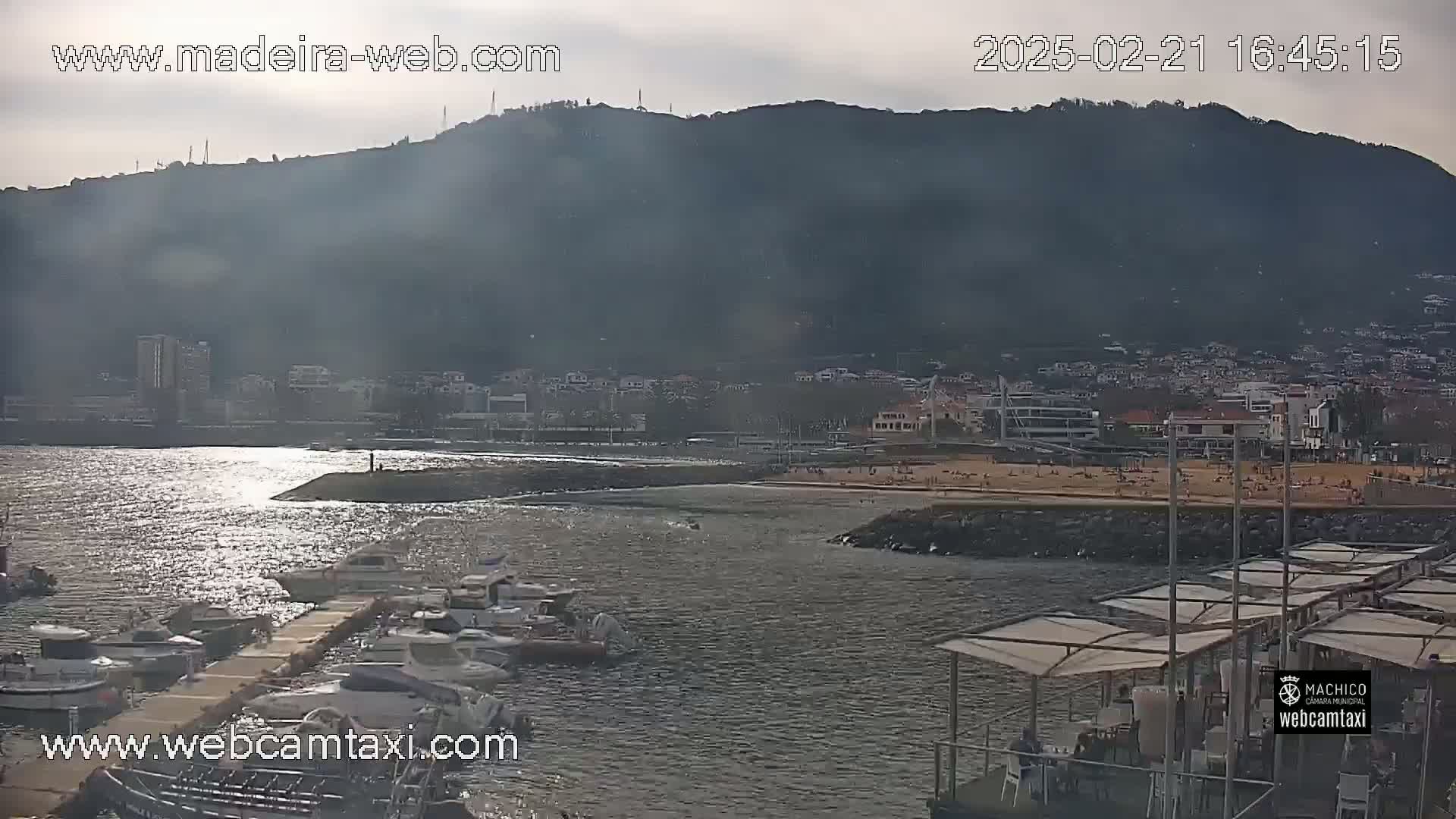 A calm body of water with boats docked at a marina, a sandy beach with people on it, and a mountain in the background under a partly cloudy sky.