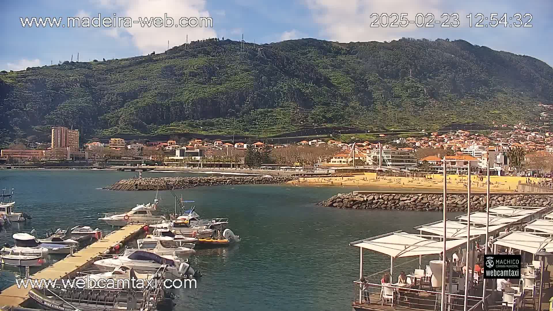 A sunny day reveals a harbor filled with boats, a sandy beach with people, and a town nestled against a verdant hillside.