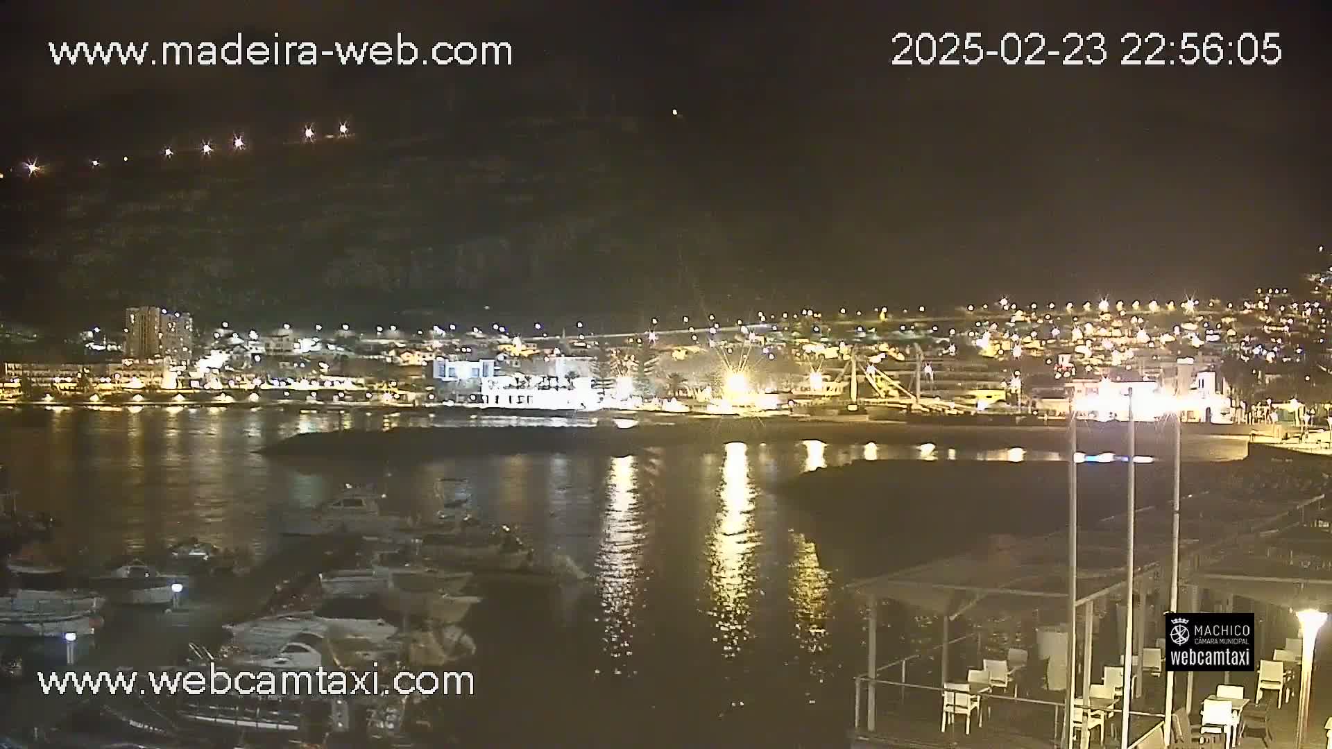A nighttime view of a harbor with many boats, calm water reflecting city lights on a hill in the background under a clear sky.