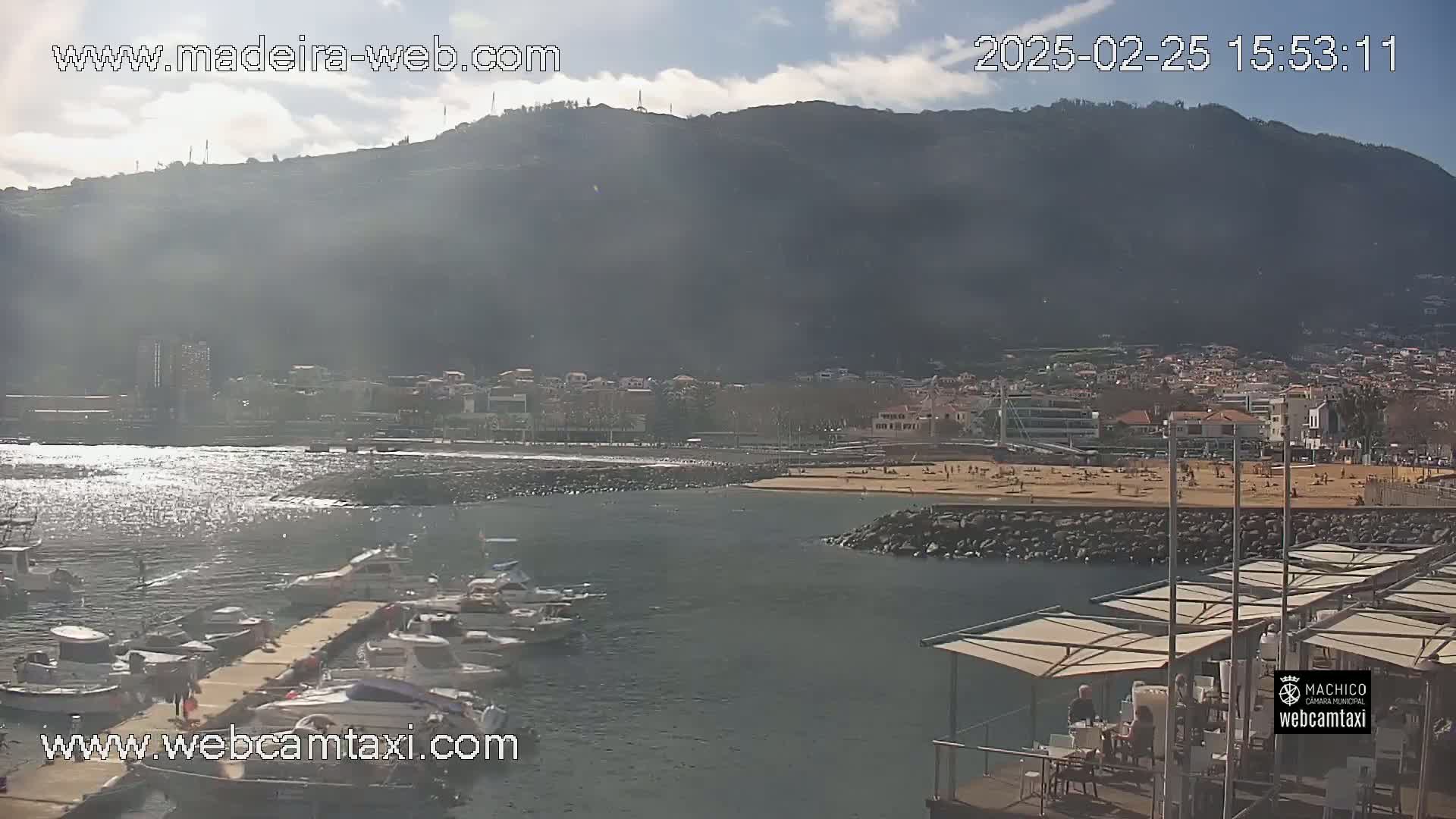 A sunny day shows a harbor with boats at a dock, a sandy beach with people, and a large mountain in the background.