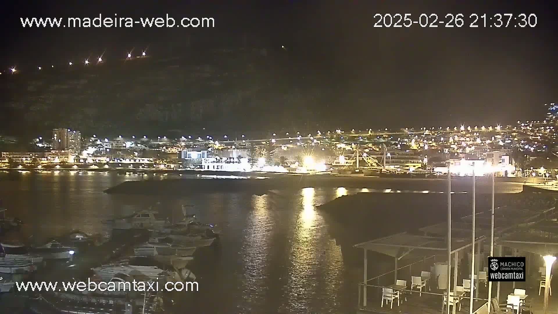 A nighttime view of a harbor with boats docked, a shoreline with lights, and a town illuminated on a hill in the background under a clear sky.