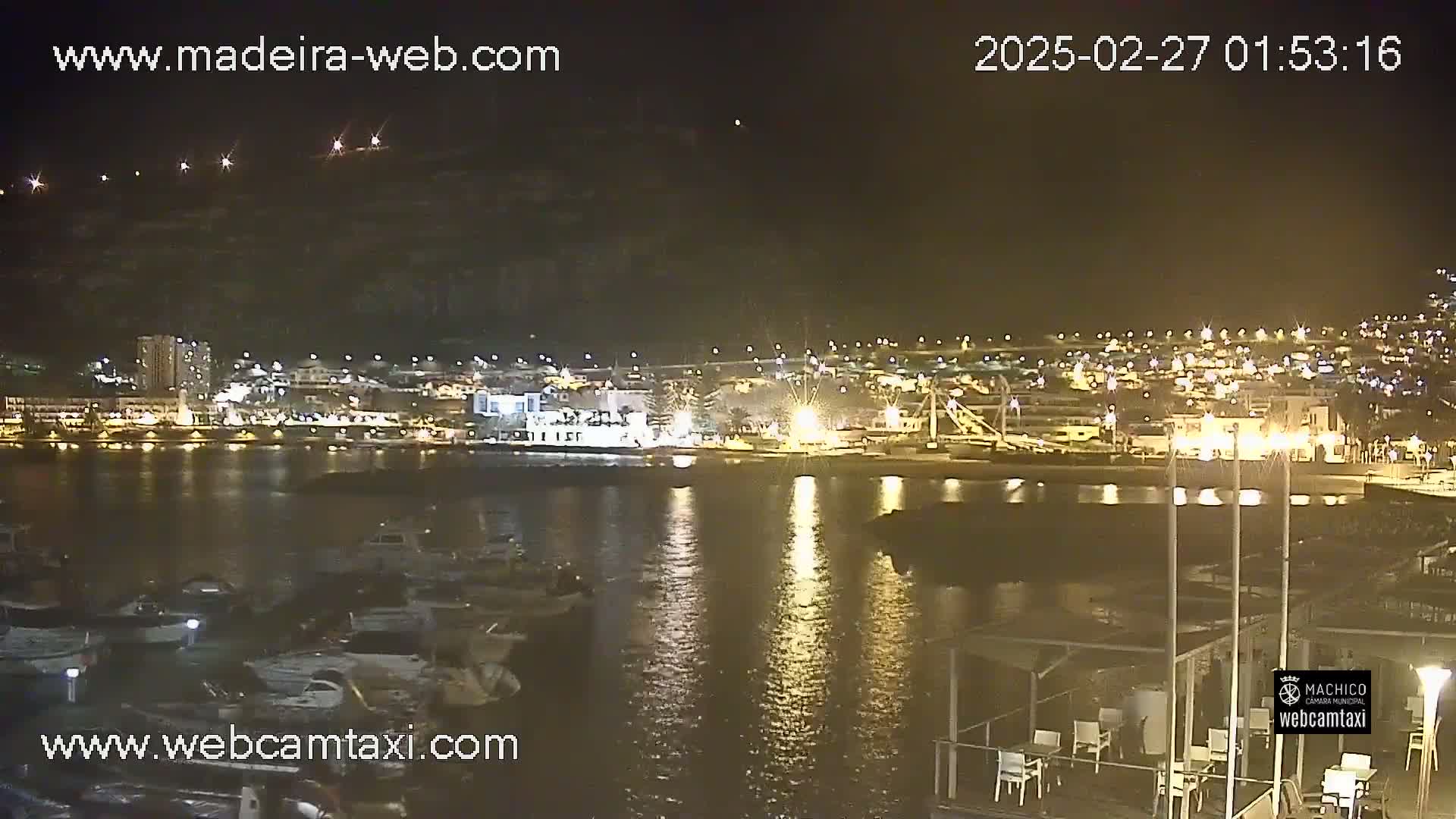 A nighttime view of a calm harbor with boats docked, reflecting lights from a city on the shore under a clear sky.
