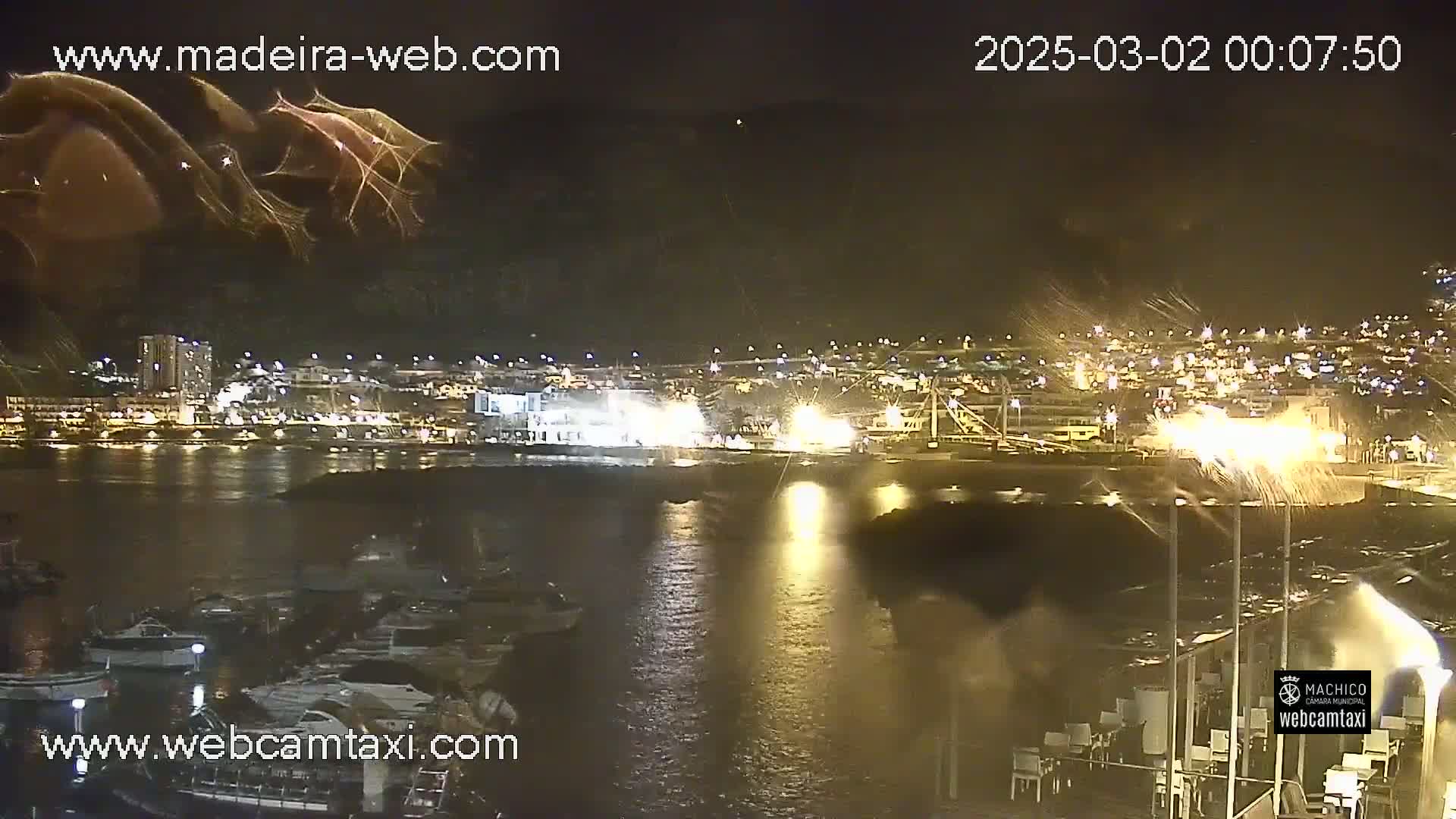 A nighttime view of a harbor town with many lights reflecting on the water, under a dark, possibly windy sky.