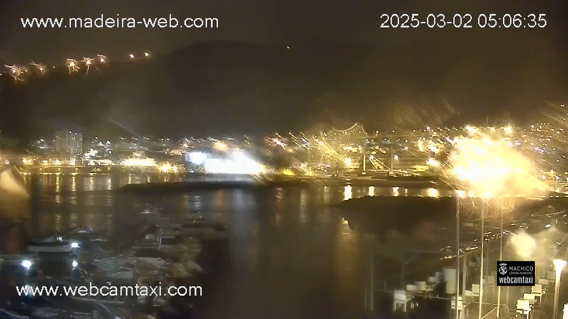 A nighttime harbor scene with fireworks exploding over a waterfront city, rain falling, and boats moored in the water.