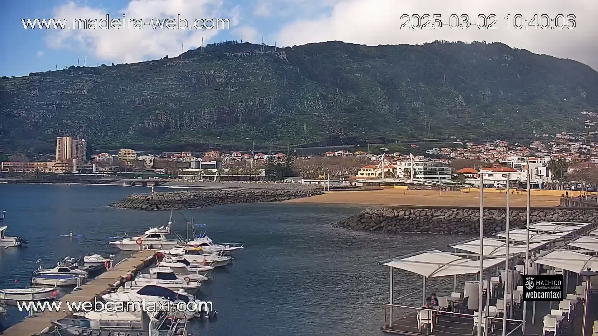 A harbor filled with boats sits at the base of a green hill under partly cloudy skies.