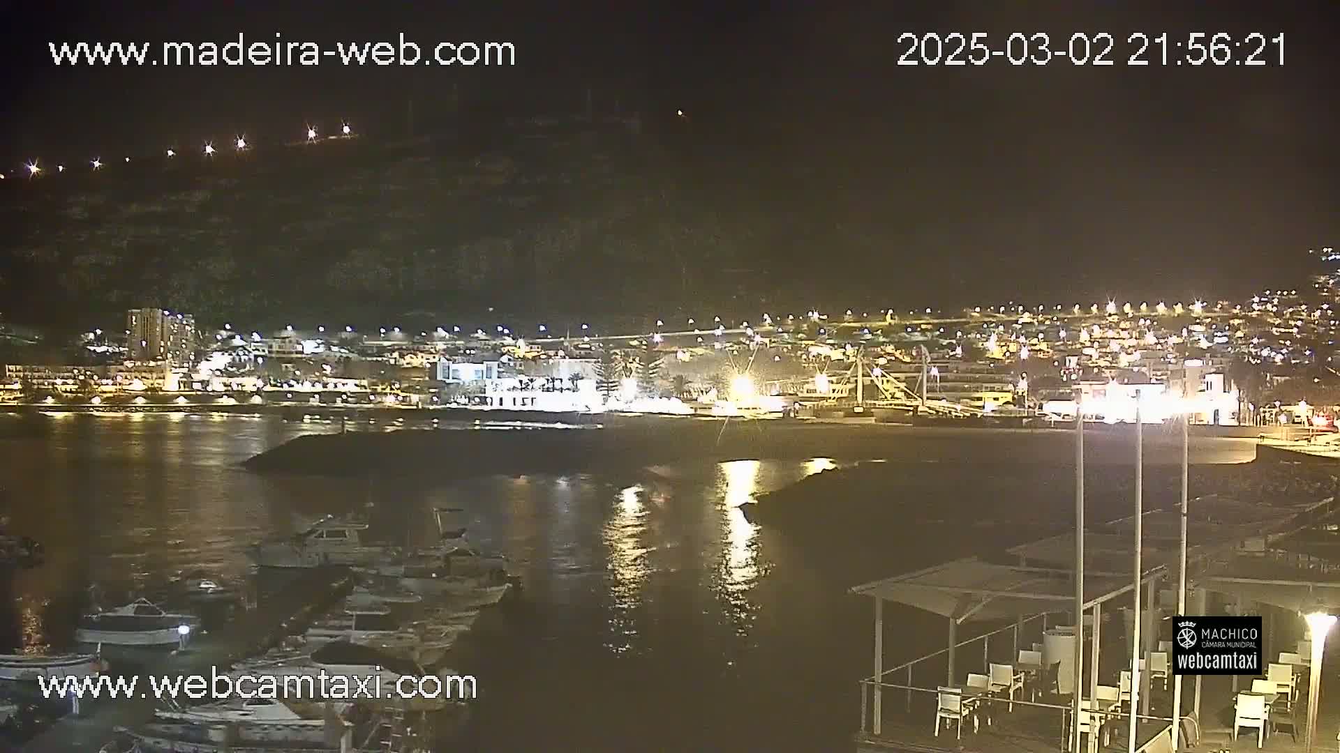 A nighttime view of a harbor filled with boats, with a town illuminated on the far shore under a clear sky.