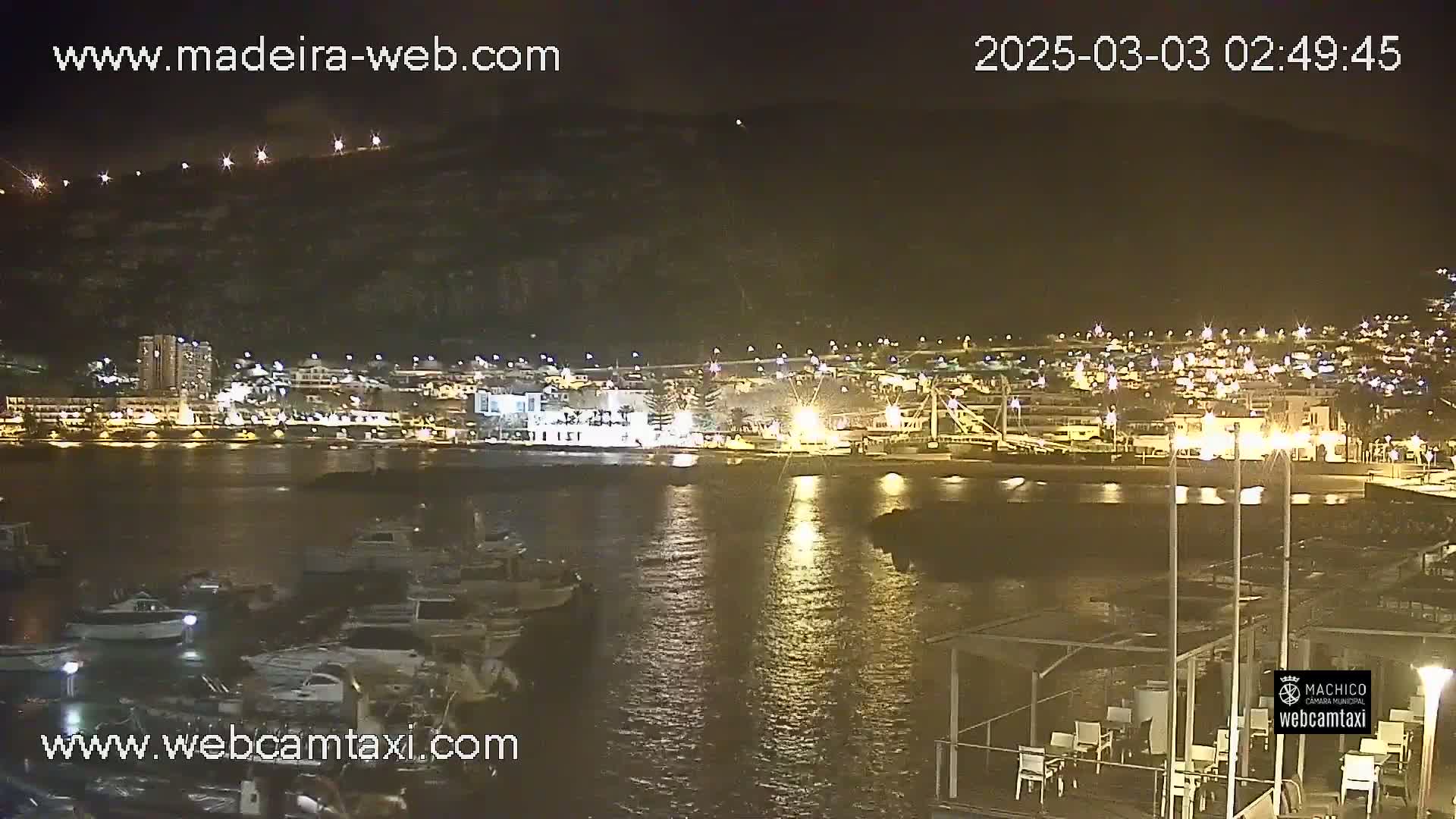 A nighttime view of a harbor filled with boats, next to a town illuminated by lights, all situated beneath a dark hill.