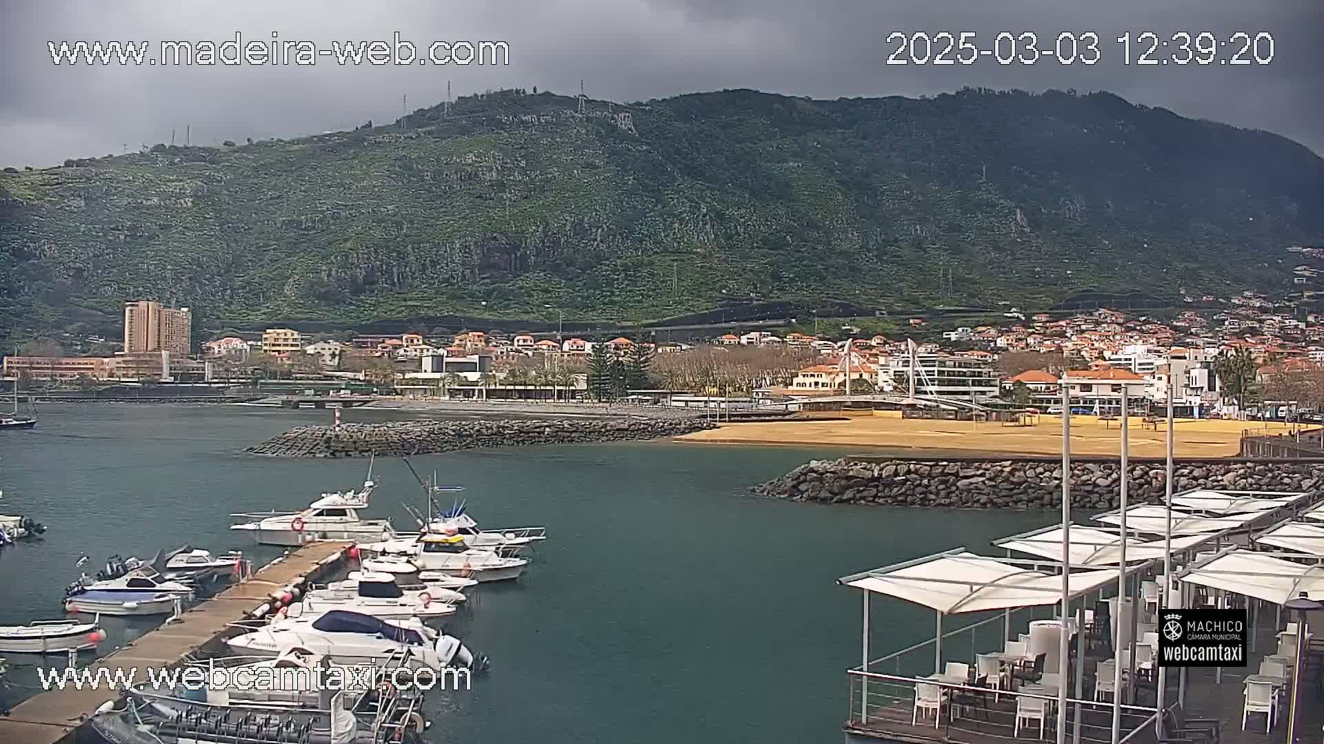 A harbor with several boats docked at a pier, under an overcast sky, is situated below a green hillside with buildings and a sandy beach in the background.