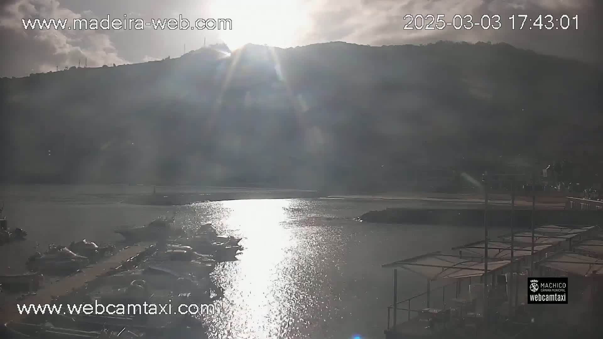 A partly cloudy sunset over a calm body of water with boats docked near a shoreline and structures with overhead coverings.