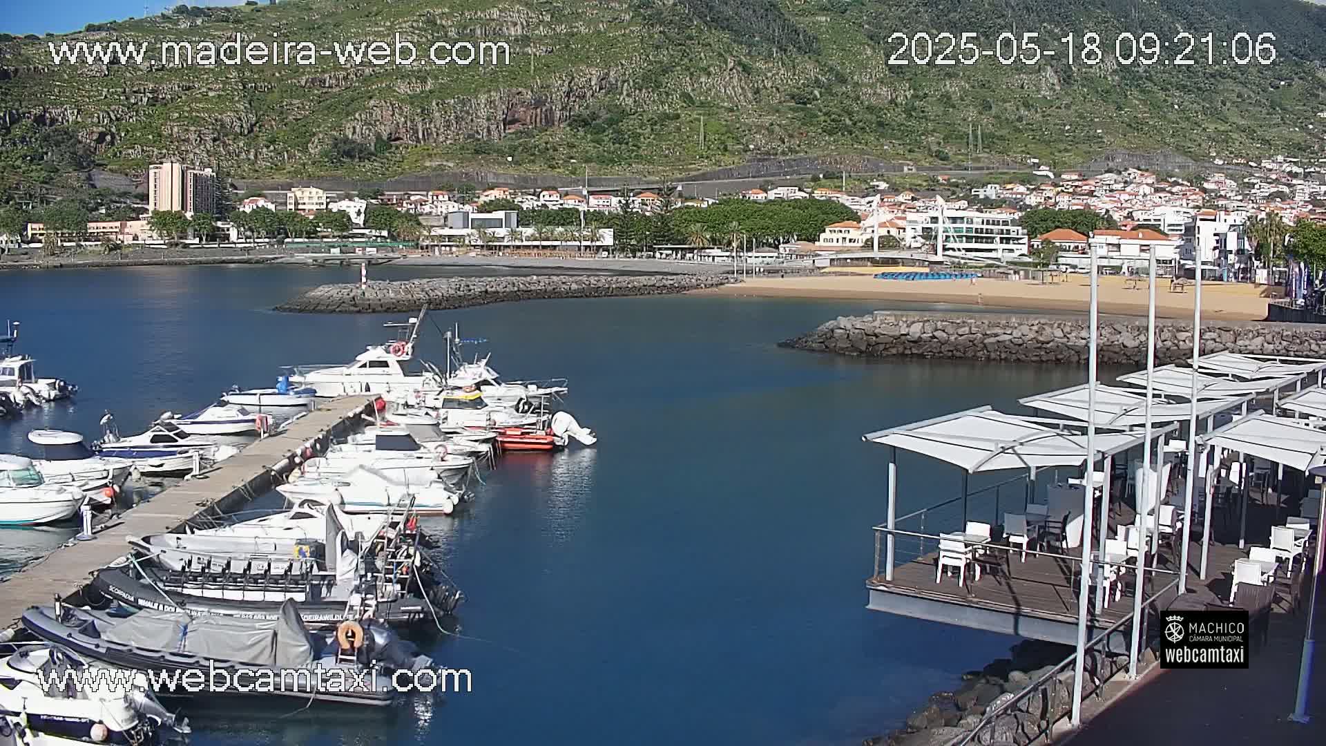 A marina filled with boats is situated next to a sandy beach and a town, all nestled against a hillside on a sunny day.
