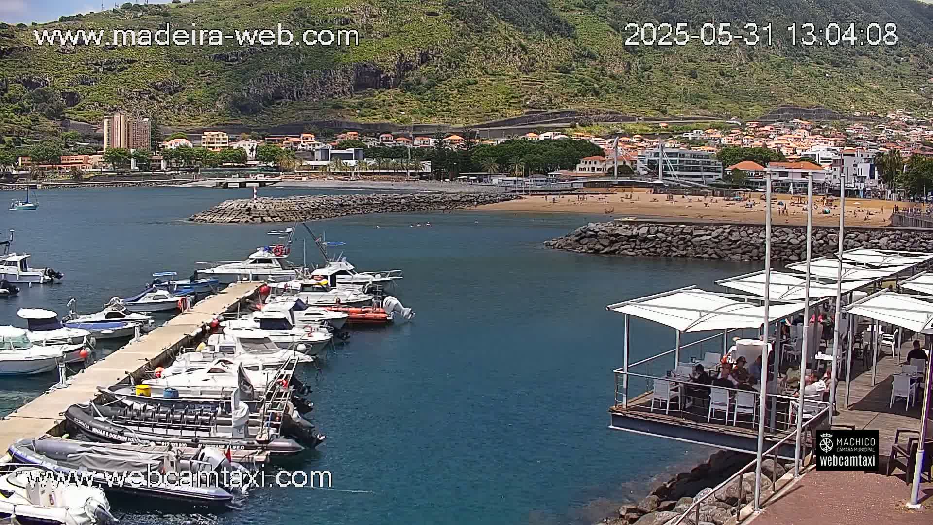 A marina filled with boats is situated next to a sandy beach and town, backed by a green mountainside under a sunny sky.