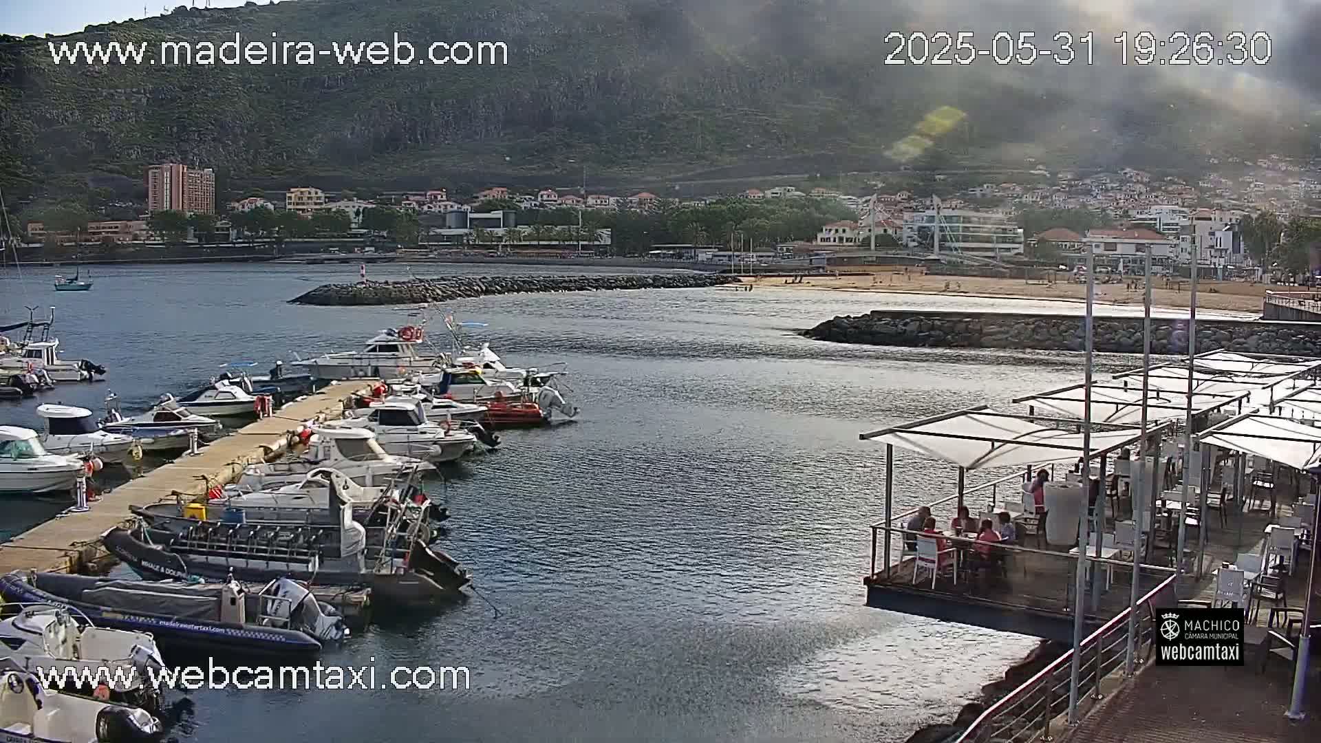 A marina filled with various sized boats is situated next to a calm body of water and a sandy beach, with buildings and hills visible in the background under a mostly clear sky.