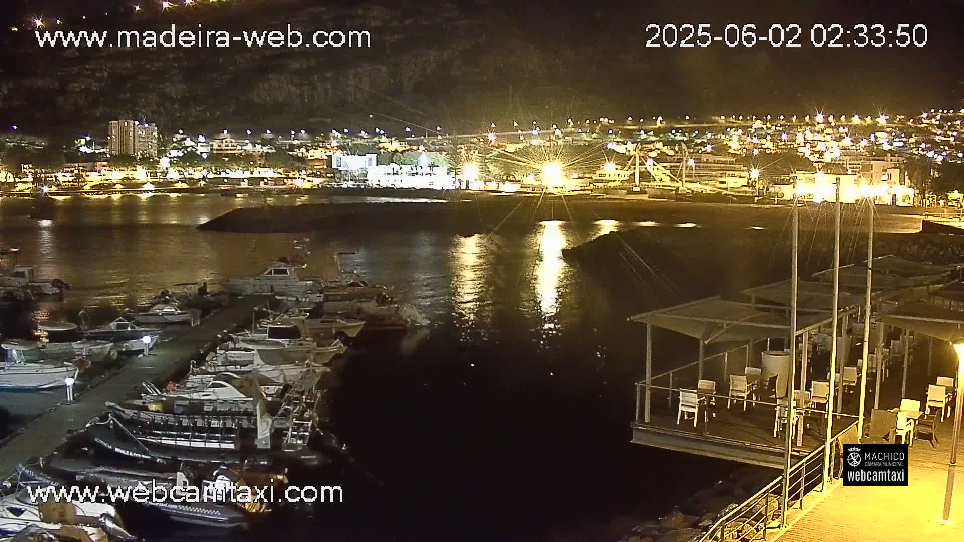 A nighttime harbor scene shows a marina filled with docked boats, a restaurant with outdoor seating on a floating dock, and a brightly lit town in the background.