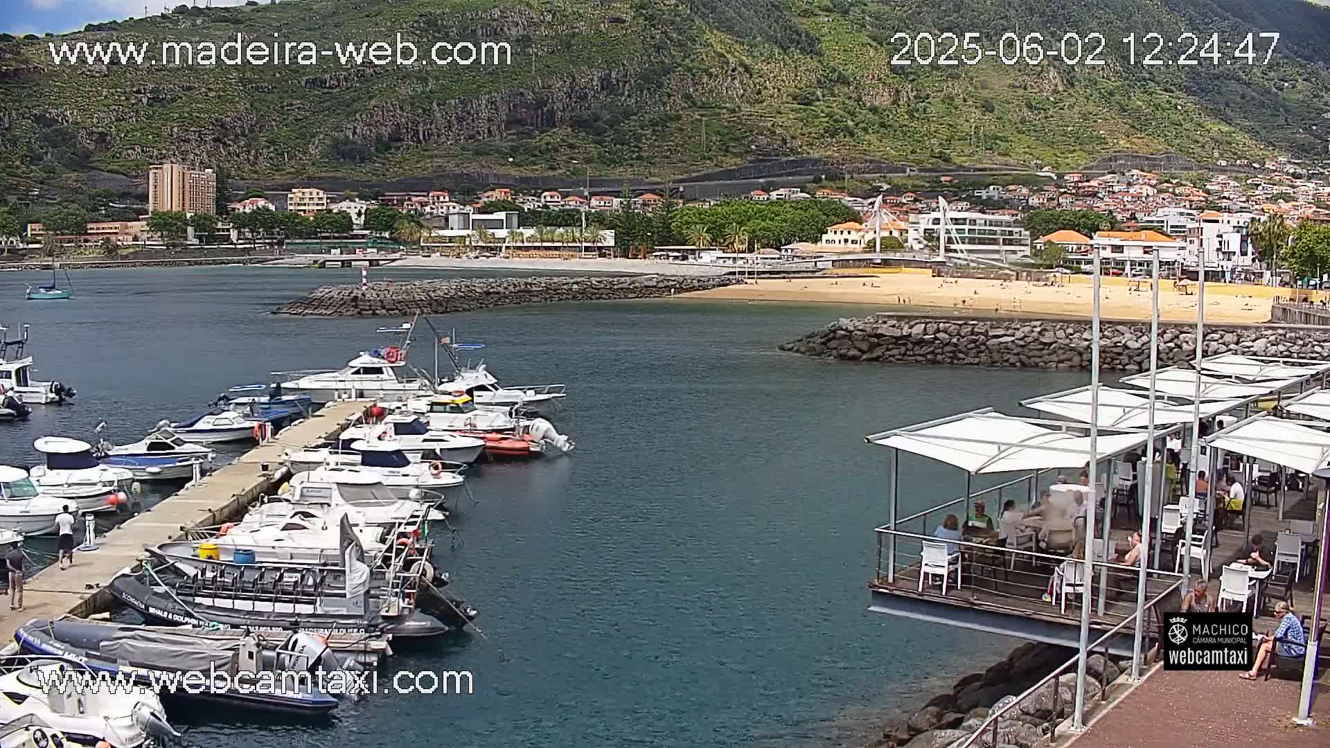 A marina filled with numerous boats is situated near a sandy beach and town, all nestled against a mountainous backdrop under sunny skies.