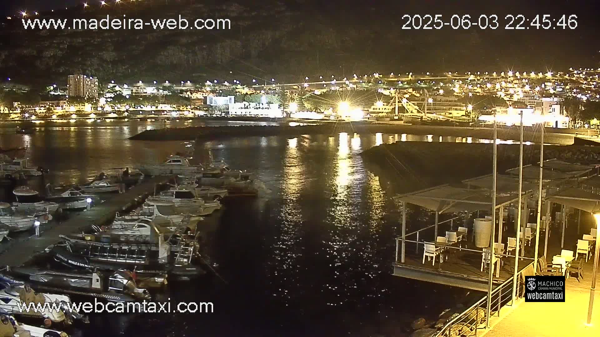 A nighttime view of a harbor filled with docked boats, adjacent to a brightly lit town situated against a dark hillside.