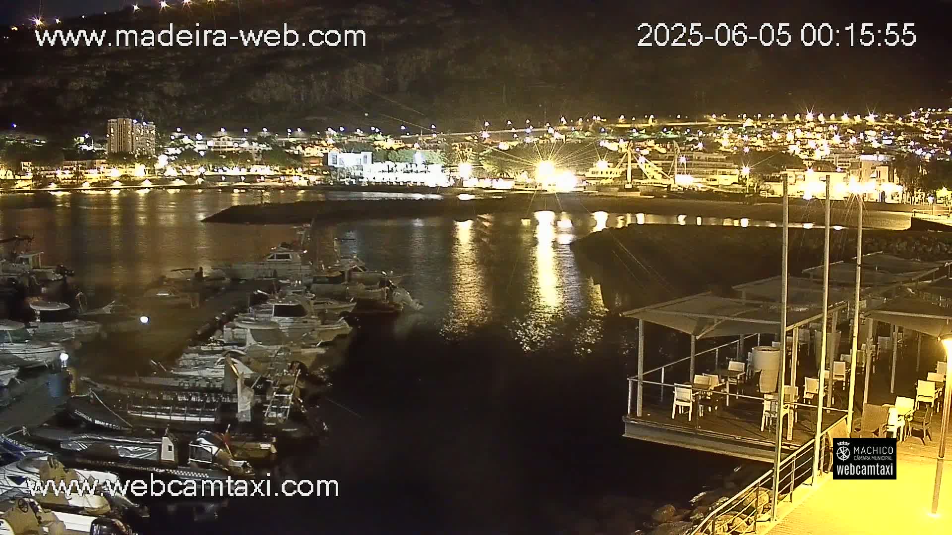 A nighttime harbor scene shows numerous boats docked, a restaurant on a floating platform, and a town illuminated in the background.