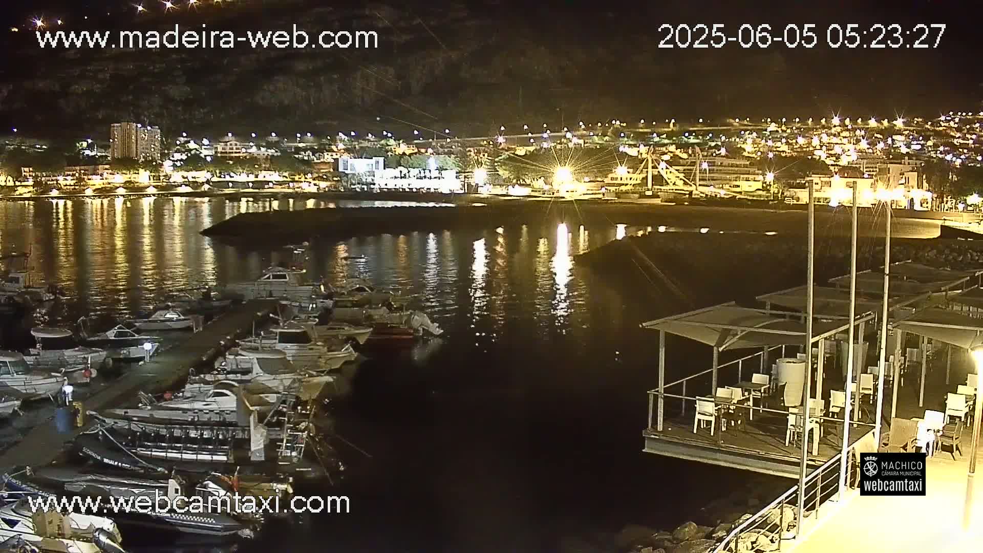 A nighttime view of a harbor filled with boats, adjacent to a lit-up town with a waterfront restaurant.