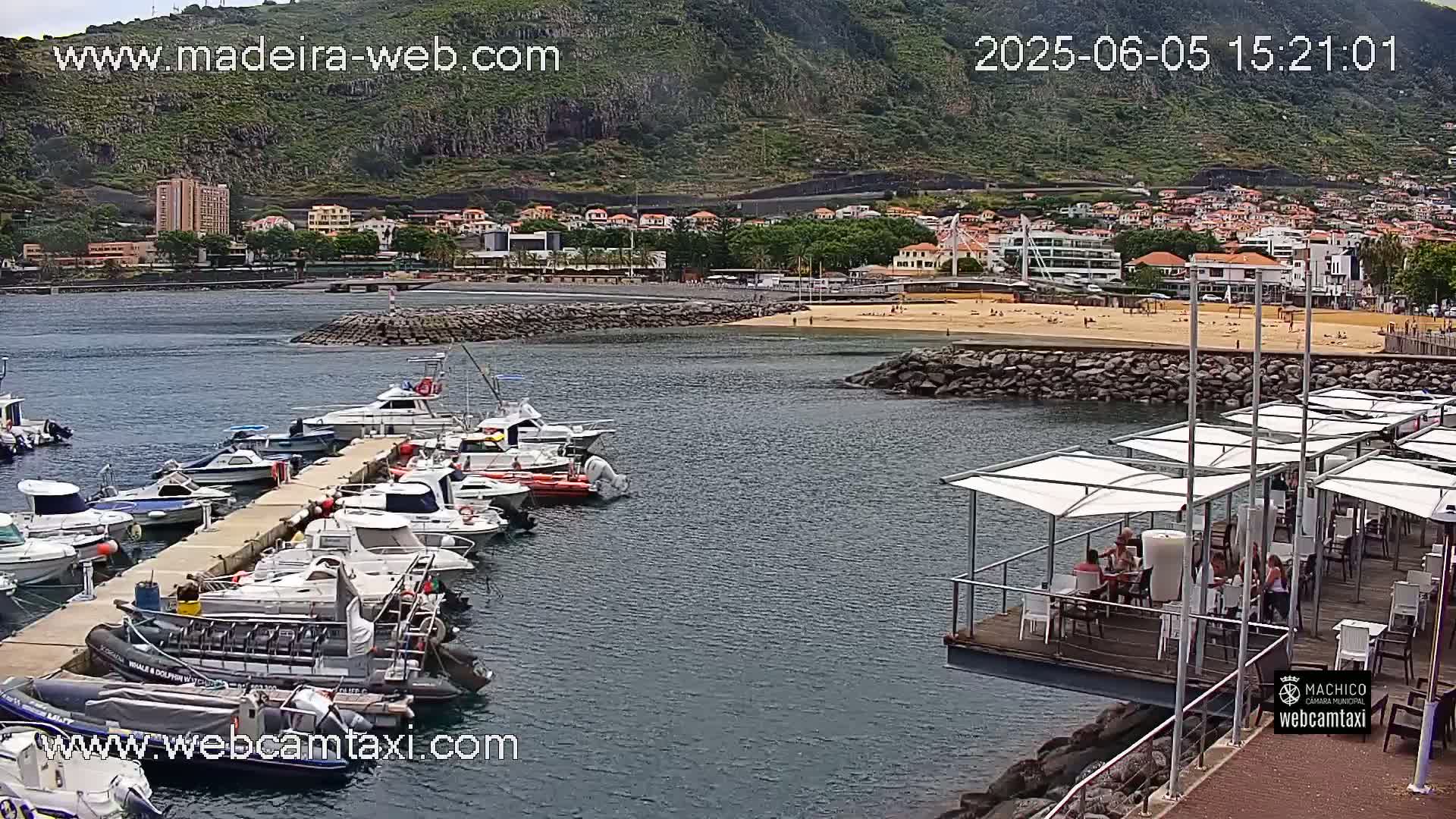 A marina filled with boats is situated next to a sandy beach and a town nestled against a green, mountainous hillside on a partly sunny day.