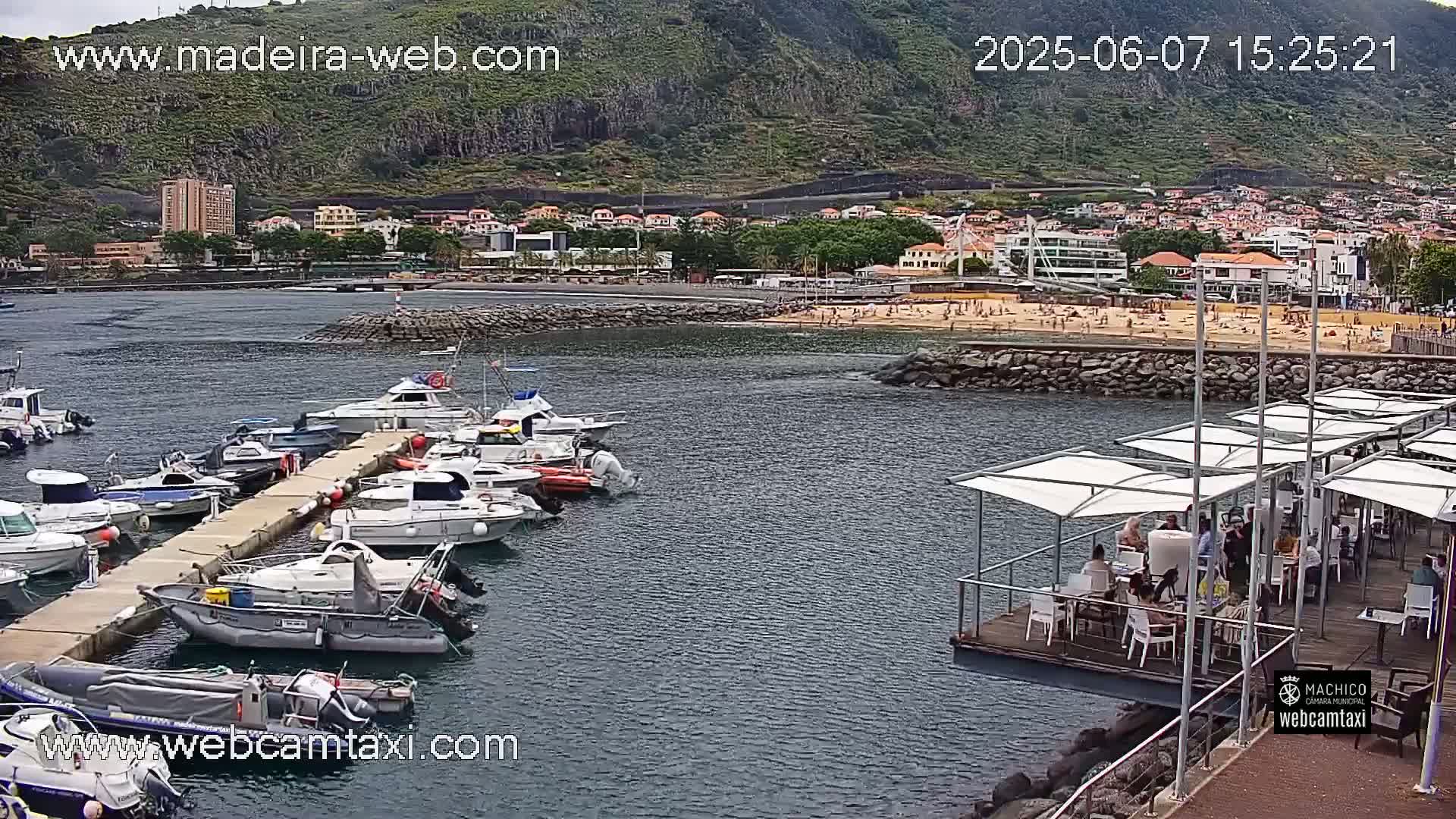 A harbor filled with numerous boats is situated near a sandy beach with people sunbathing, all set against the backdrop of a mountainous coastline under partly cloudy skies.