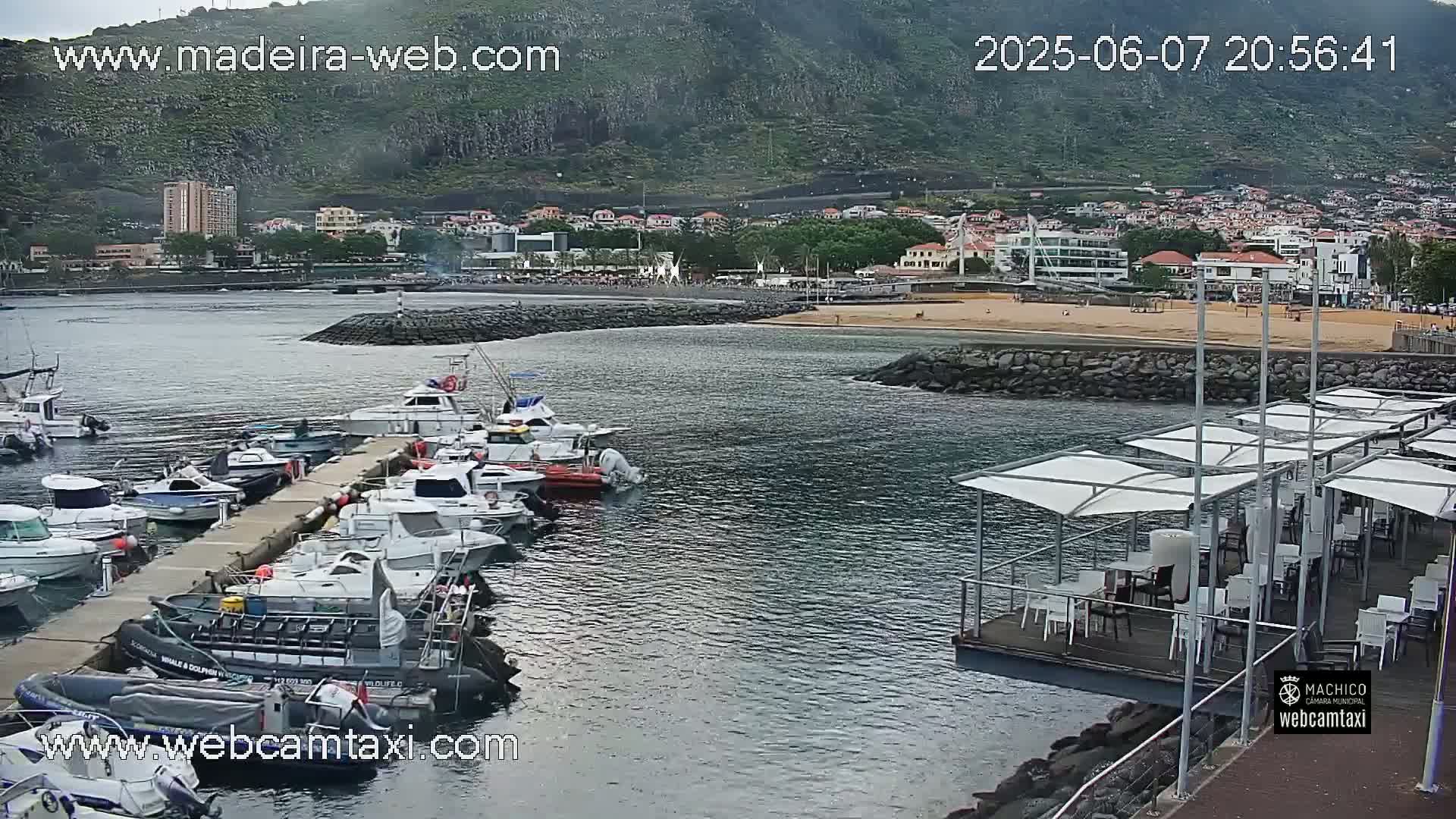 A harbor filled with docked boats sits near a sandy beach and town under an overcast sky, backed by a large, green mountain.