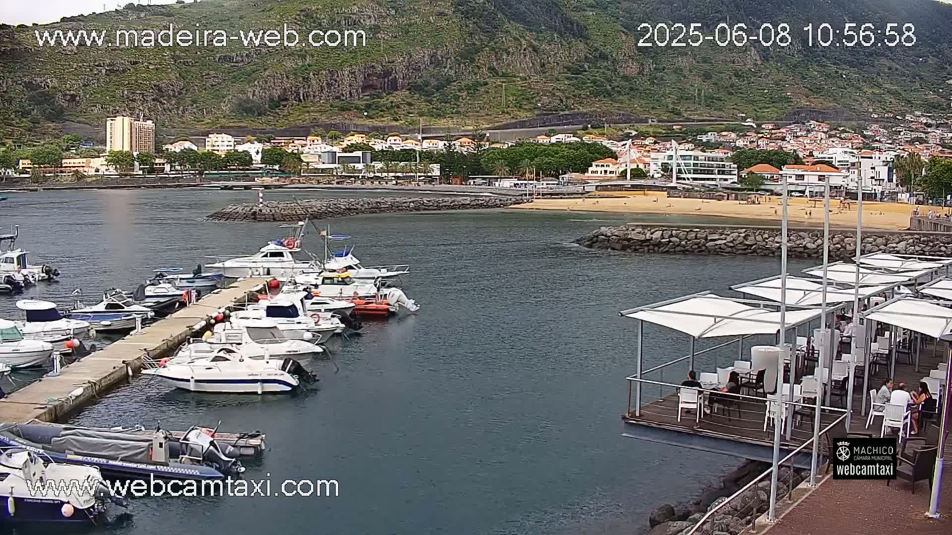 A marina filled with numerous boats is situated next to a beach and a town, all backed by a steep, green hillside under a partly cloudy sky.