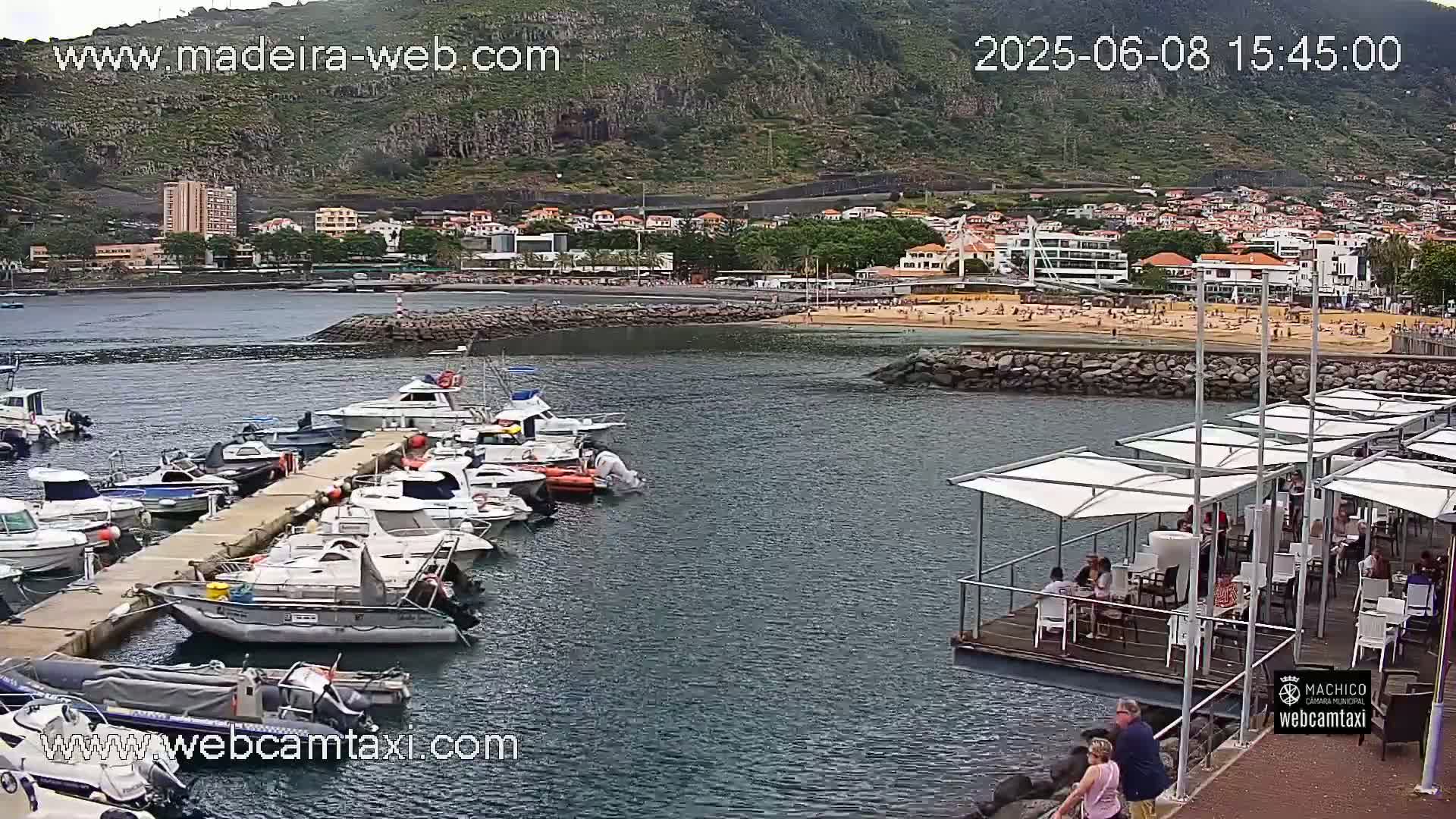 A harbor filled with numerous boats is situated near a sandy beach and a town built along a hillside under an overcast sky.