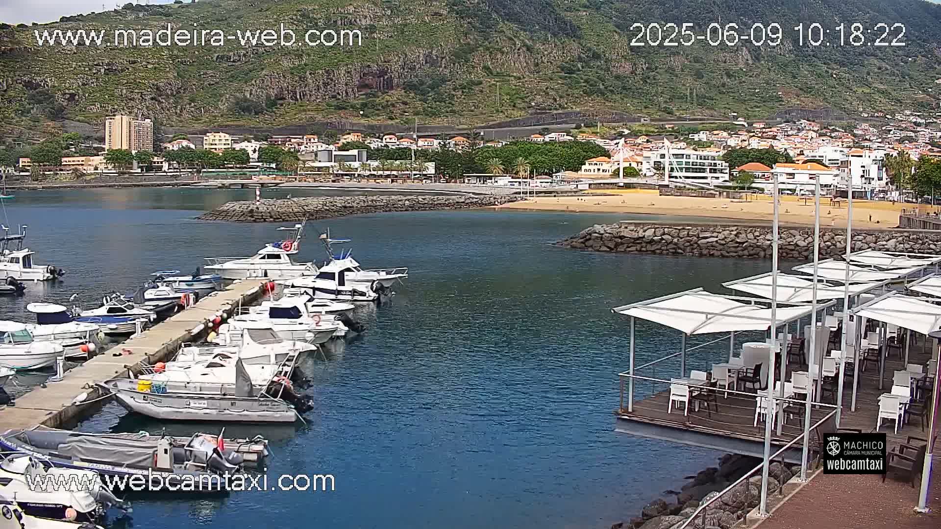 A marina filled with boats is situated in front of a sandy beach and a town nestled against a mountainous backdrop under partly cloudy skies.