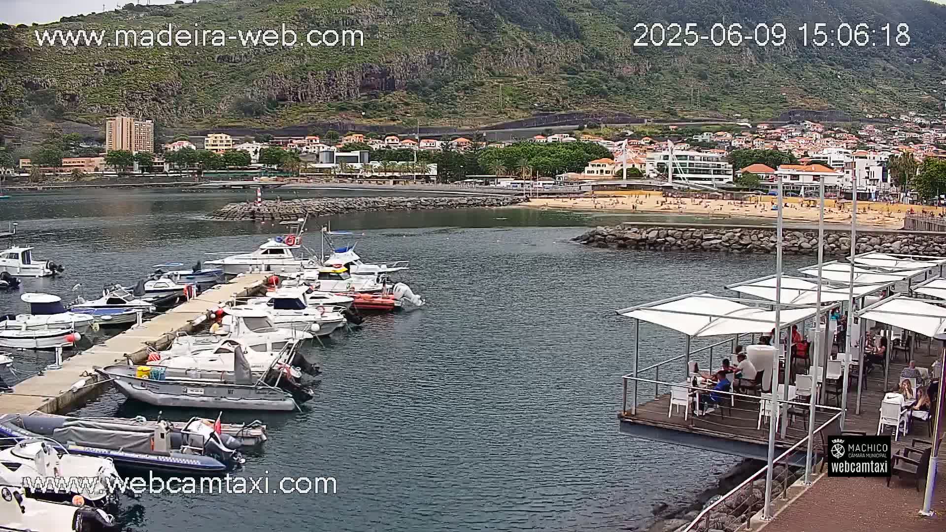 A harbor filled with docked boats is situated below a hillside town on a partly cloudy day.