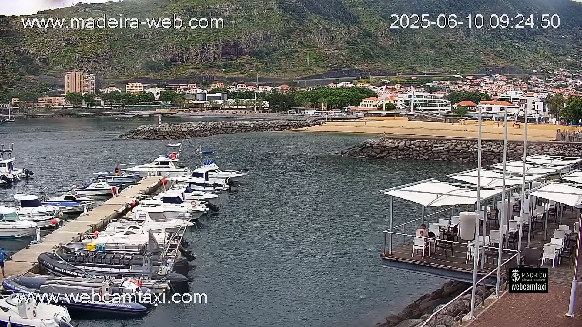 A marina filled with numerous boats is situated near a sandy beach and a town nestled against a mountainous backdrop under overcast skies.