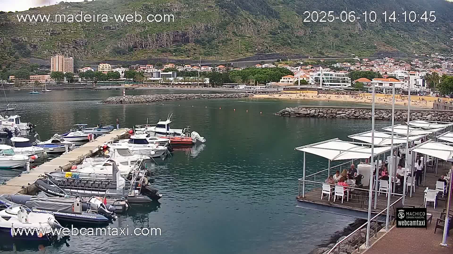 A marina filled with boats is situated near a beach with people sunbathing, all in front of a town nestled against a hillside.