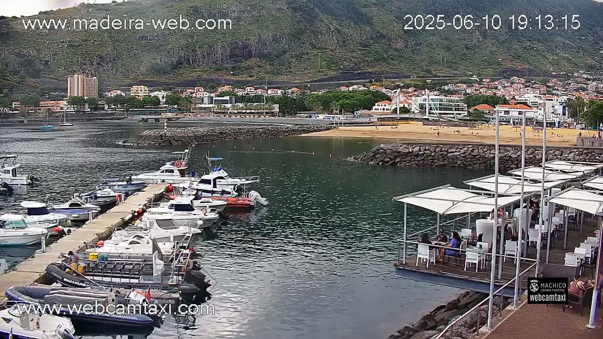 A harbor filled with boats is situated next to a sandy beach and a town at the foot of a mountain under a partly cloudy sky.
