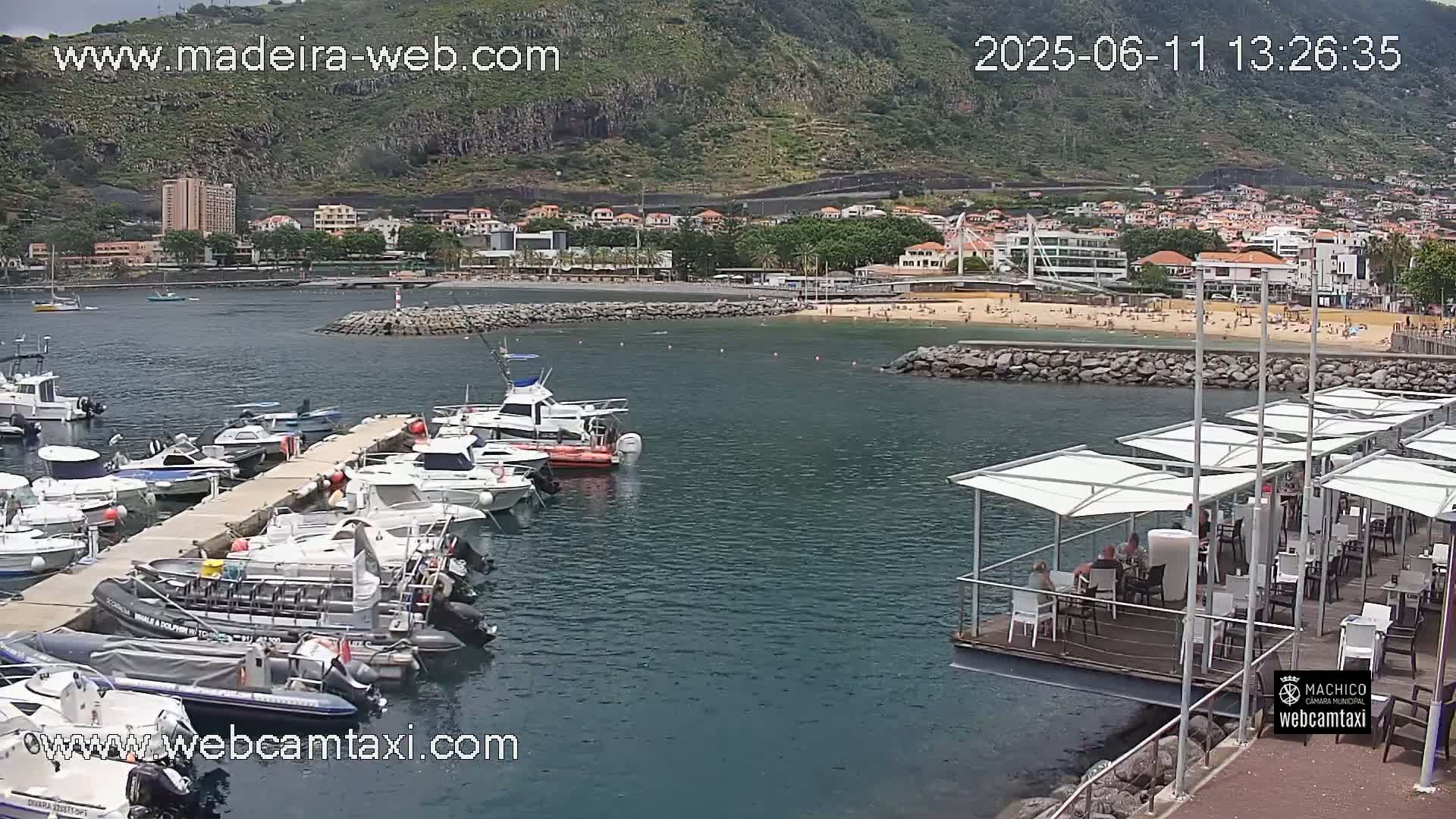A marina filled with numerous boats is situated near a beach with people sunbathing, all set against a backdrop of a mountainous town under a partly cloudy sky.