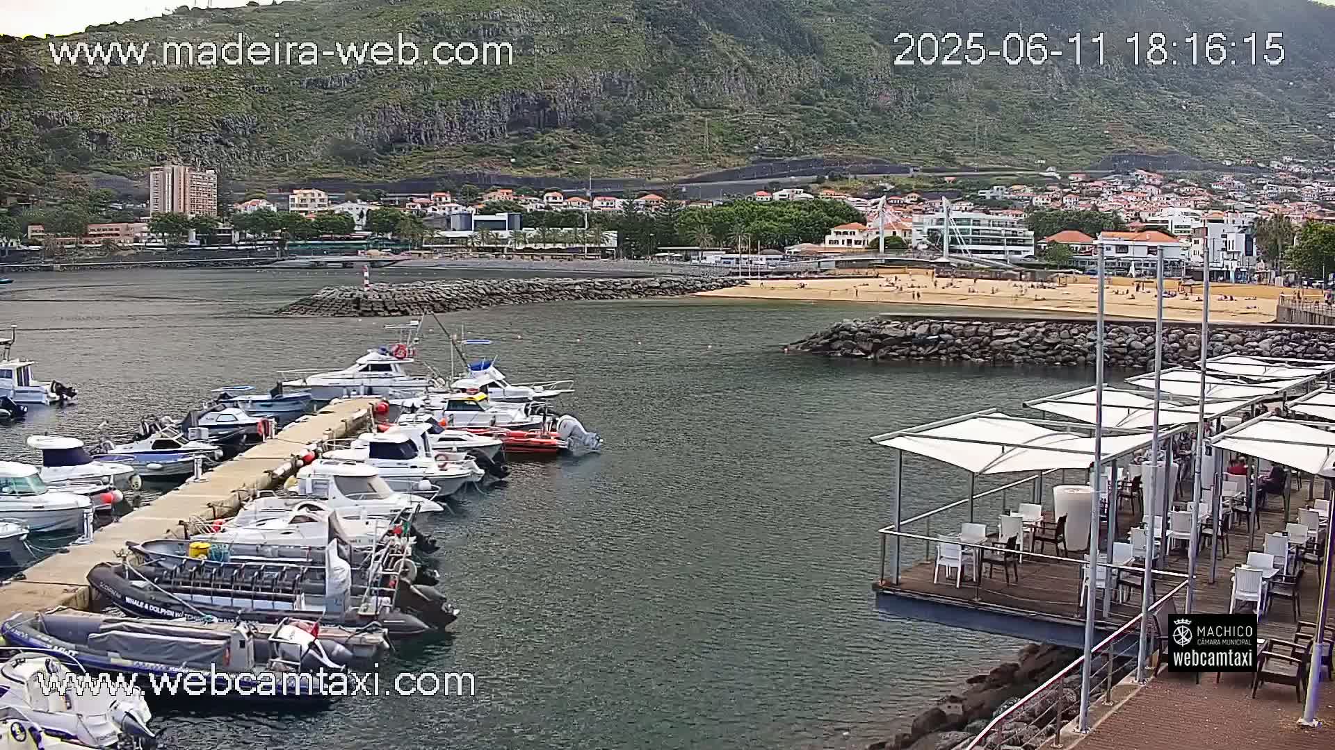 A marina filled with numerous boats is situated next to a sandy beach and a small town nestled against a mountainous backdrop under partly cloudy skies.
