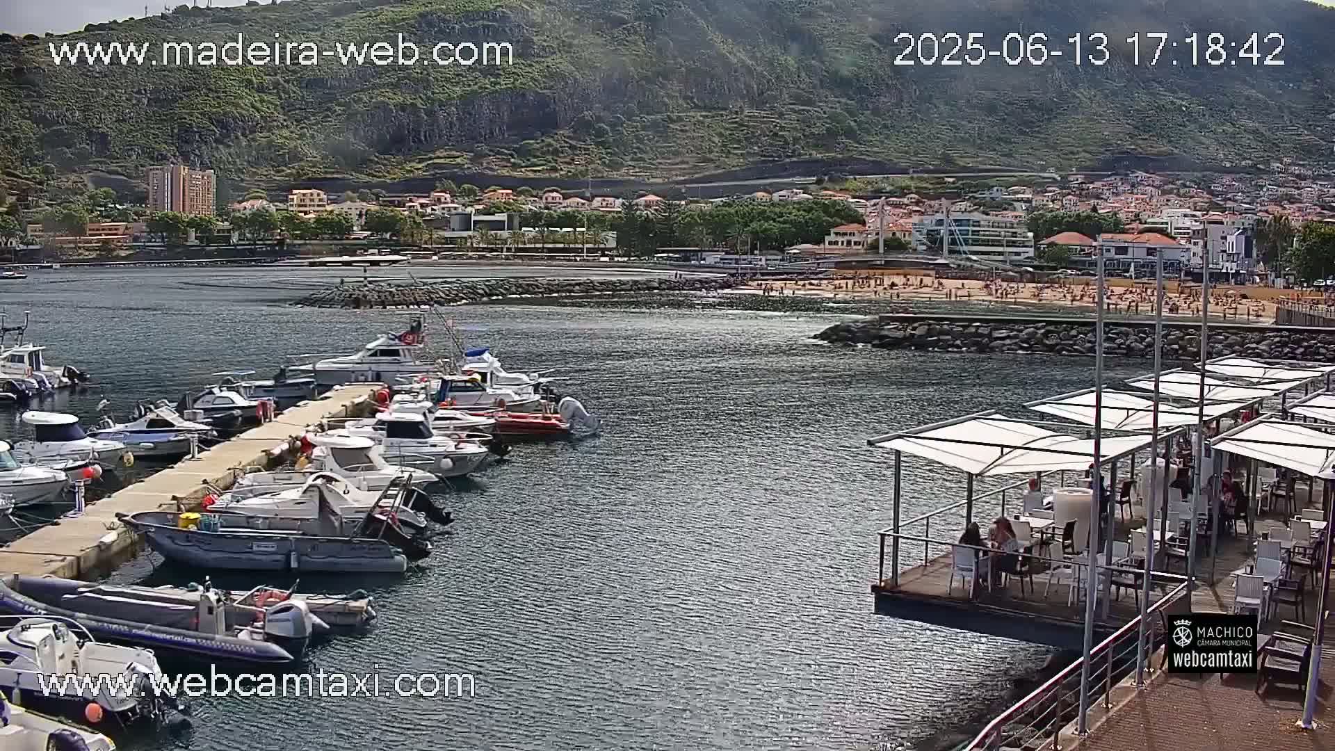 A marina filled with numerous boats is situated near a beach with many people on a sunny day, backed by a town nestled against a mountainous landscape.