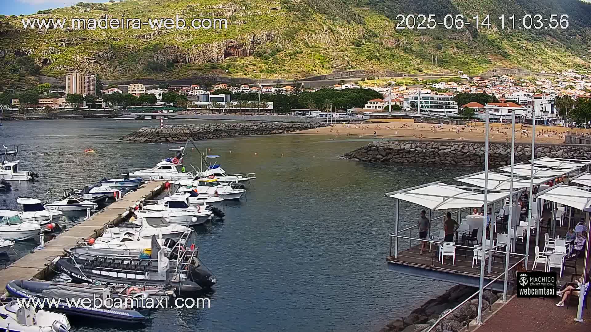 A marina filled with boats is situated next to a sandy beach and a town, with a mountainous backdrop on a sunny day.