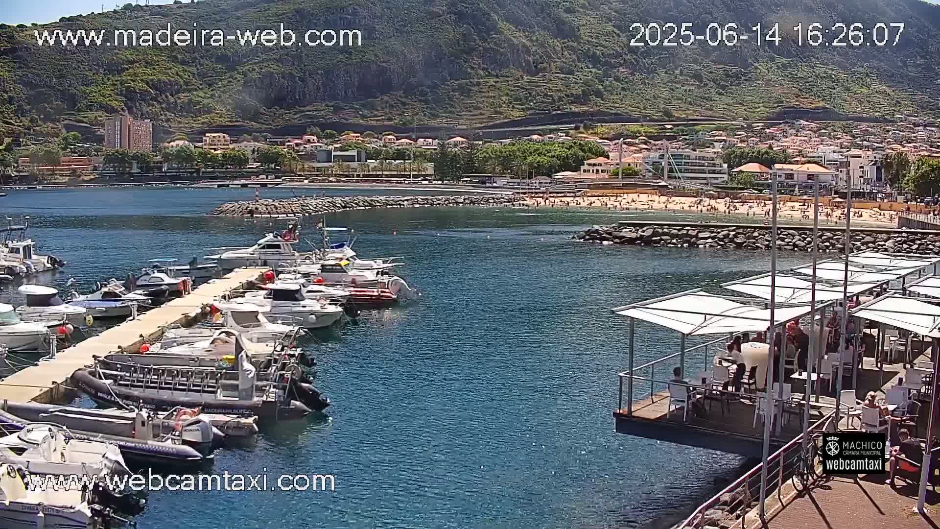 A harbor filled with boats is situated next to a sandy beach with many people on a sunny day, with a mountain in the background.