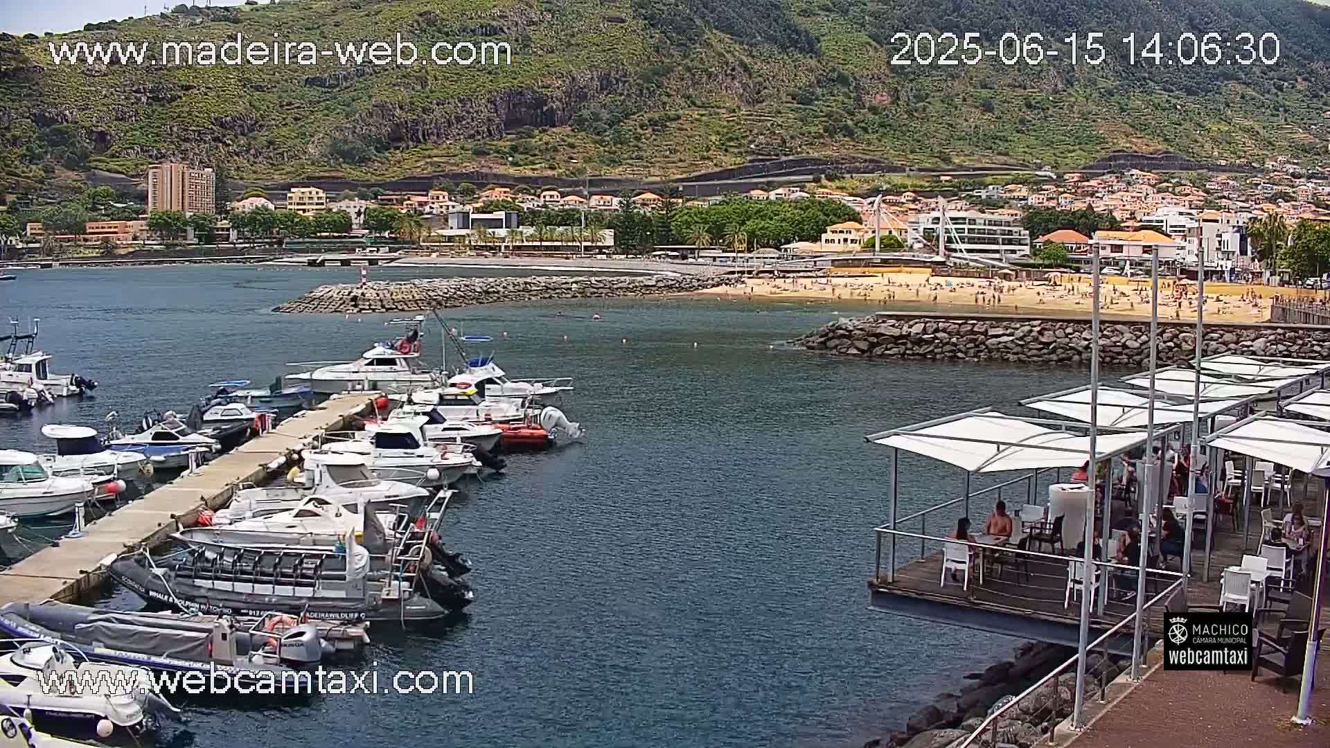 A marina filled with numerous boats is situated near a sandy beach with people sunbathing, all set against the backdrop of a hilly town under a clear sky.