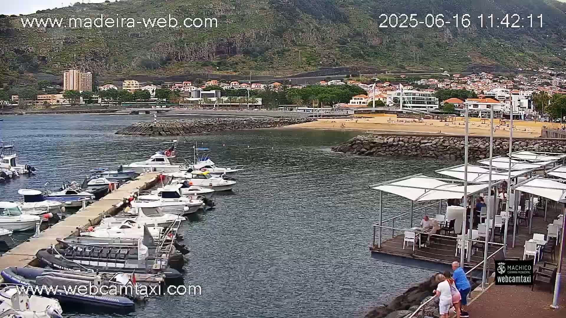 A harbor filled with numerous boats is nestled beside a sandy beach and a small town, situated against a backdrop of a mountainous region under partly cloudy skies.