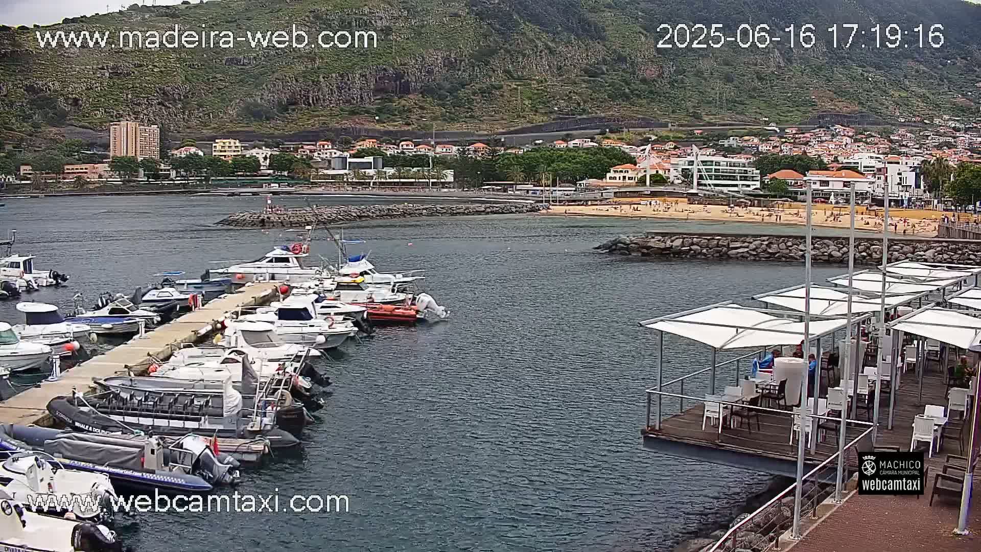 A marina filled with numerous boats is situated in front of a beach and town, backed by a large hill, under an overcast sky.